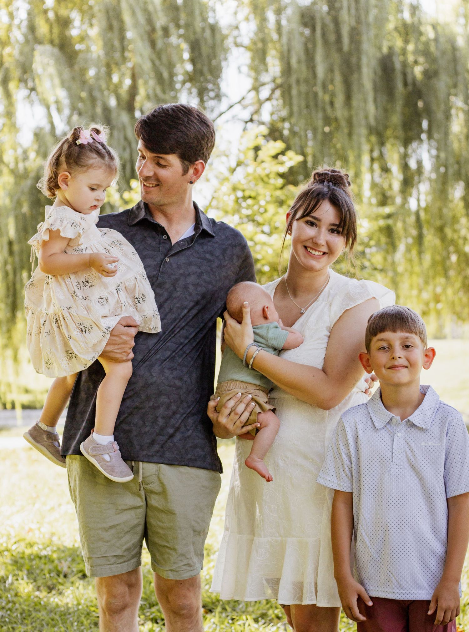 Happy family of five posing outdoors near willow trees, parents holding young children including a newborn baby.