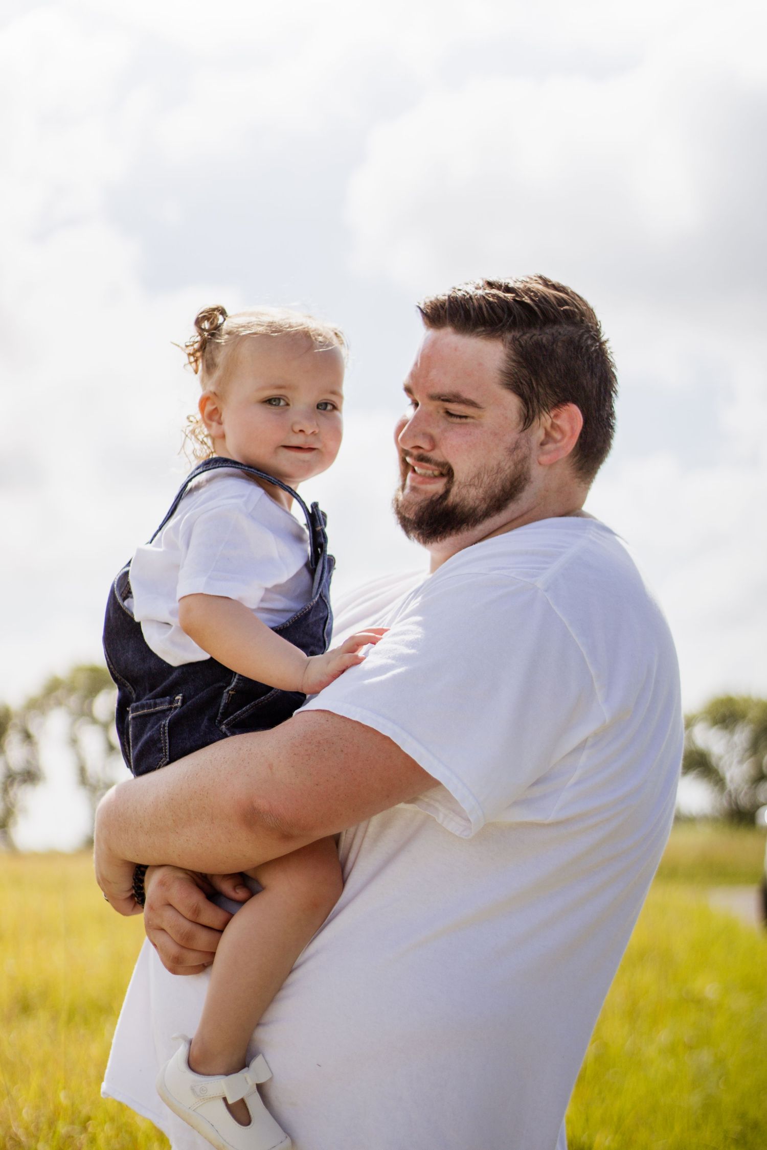 Bearded father in white t-shirt holding smiling toddler girl in denim overalls in a sunny green field.