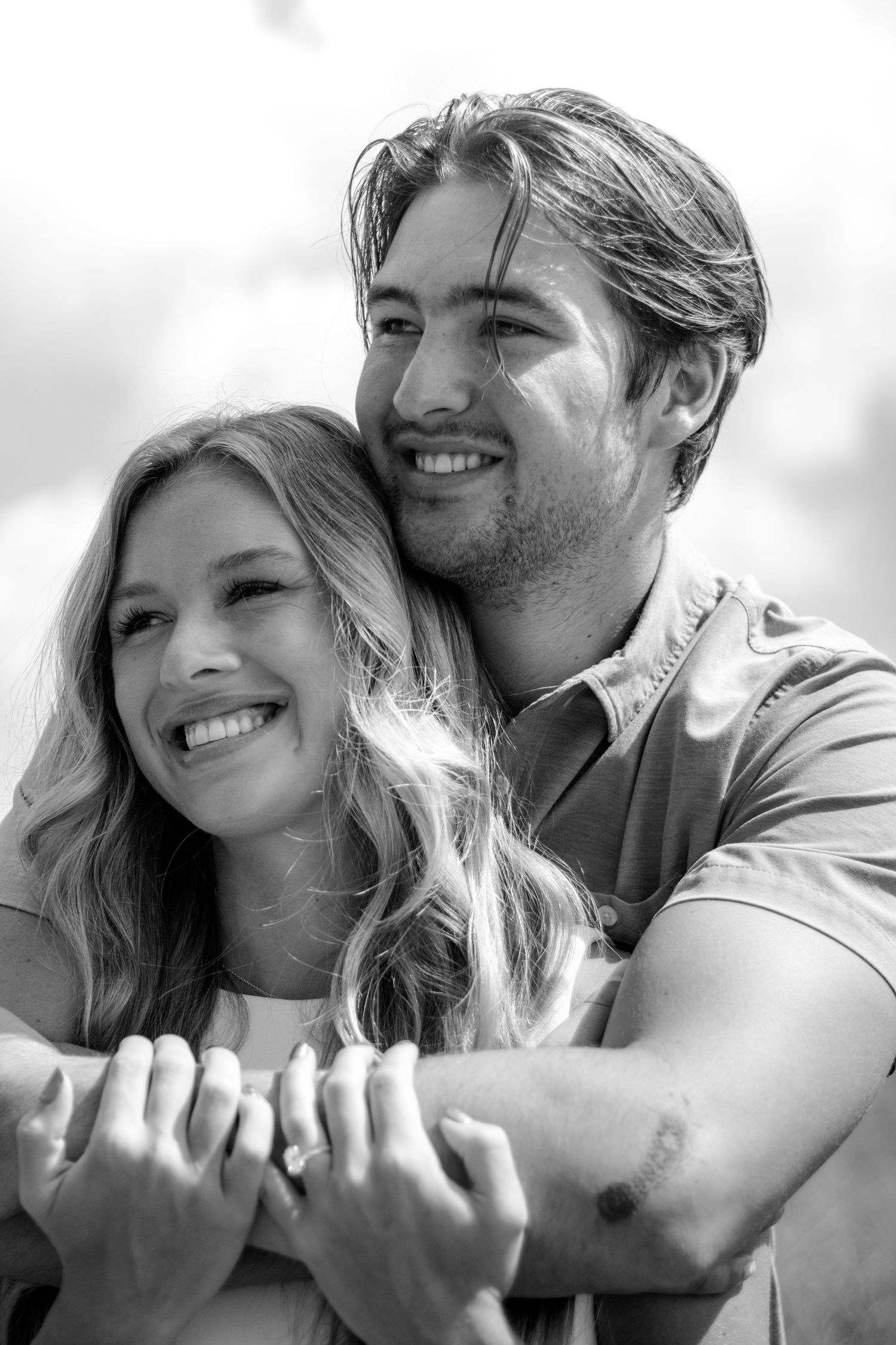 Black and white photo of a smiling couple embracing, man hugging woman from behind outdoors.