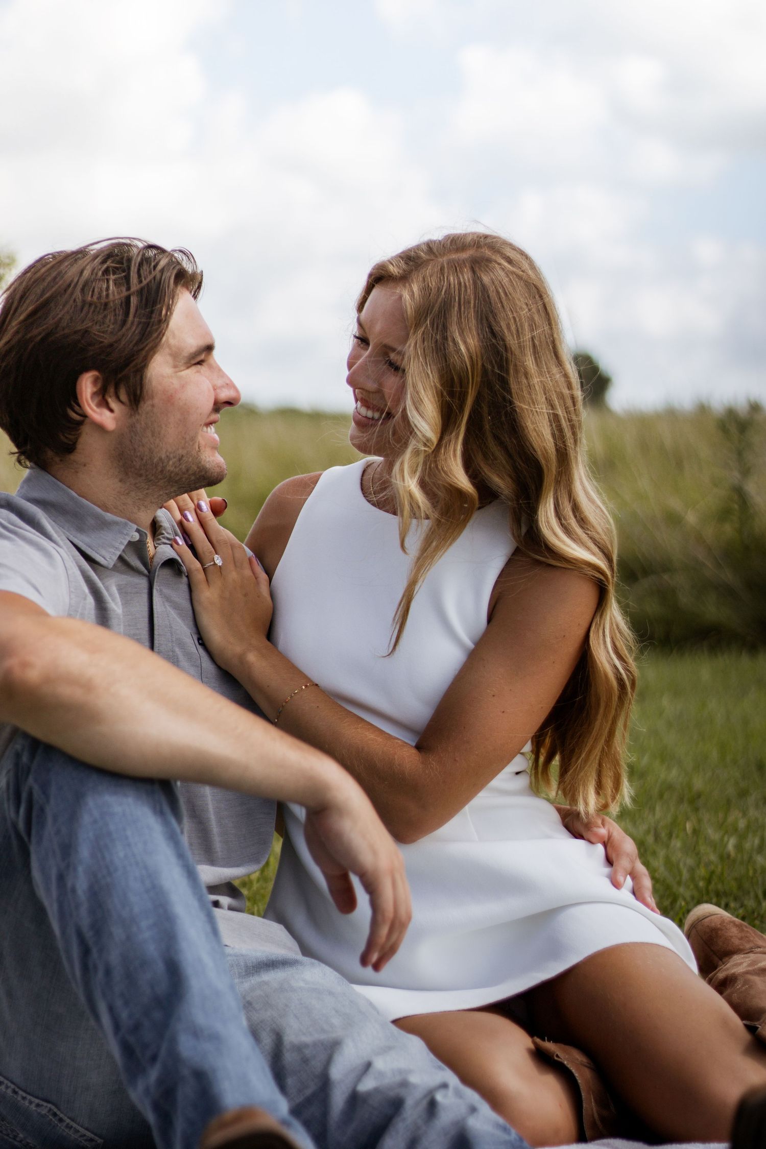 A happy couple sits together in a grassy field, smiling at each other, woman in white dress, man in jeans.