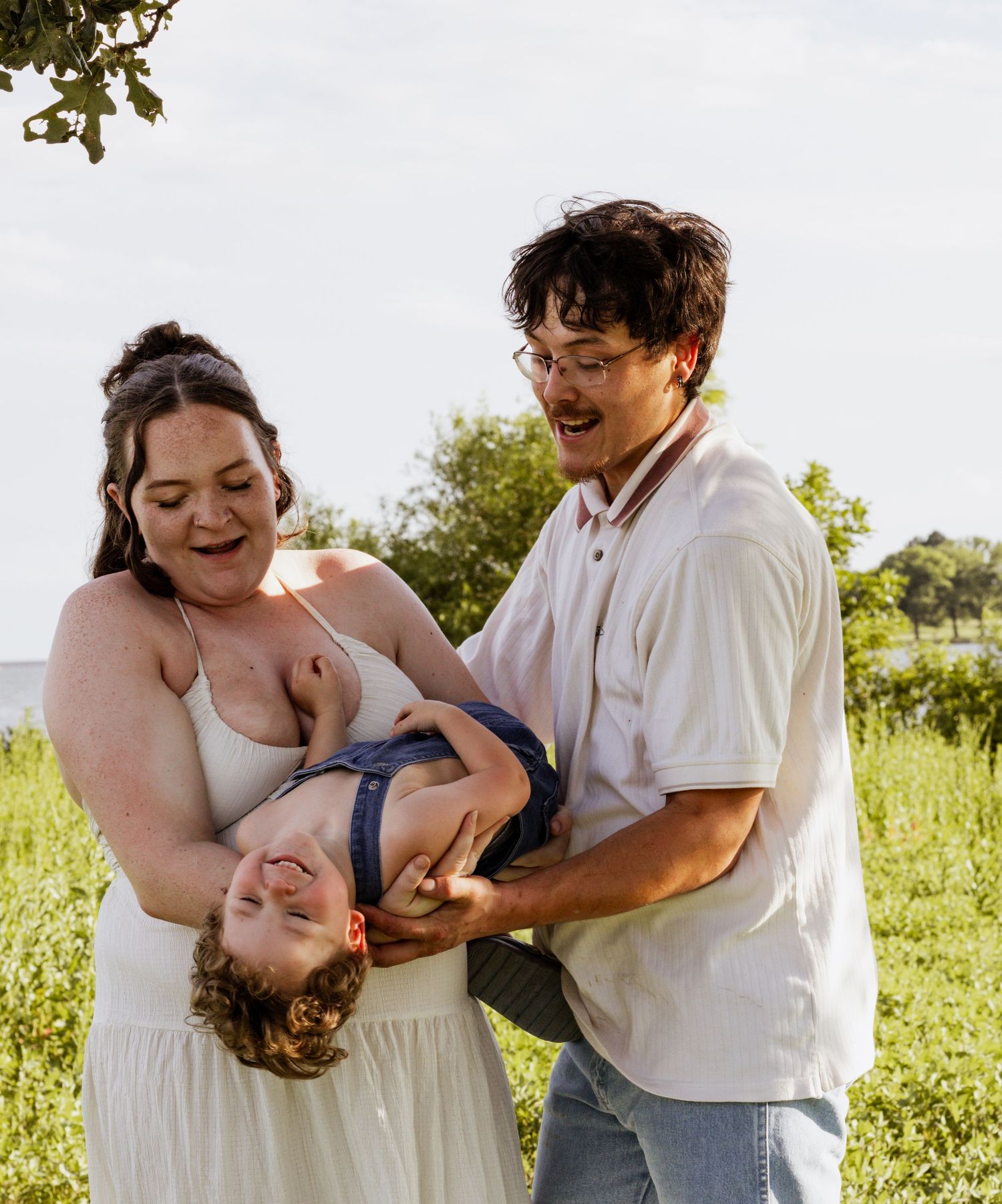 Two parents laughing while playfully holding their toddler upside down outdoors in a green field.