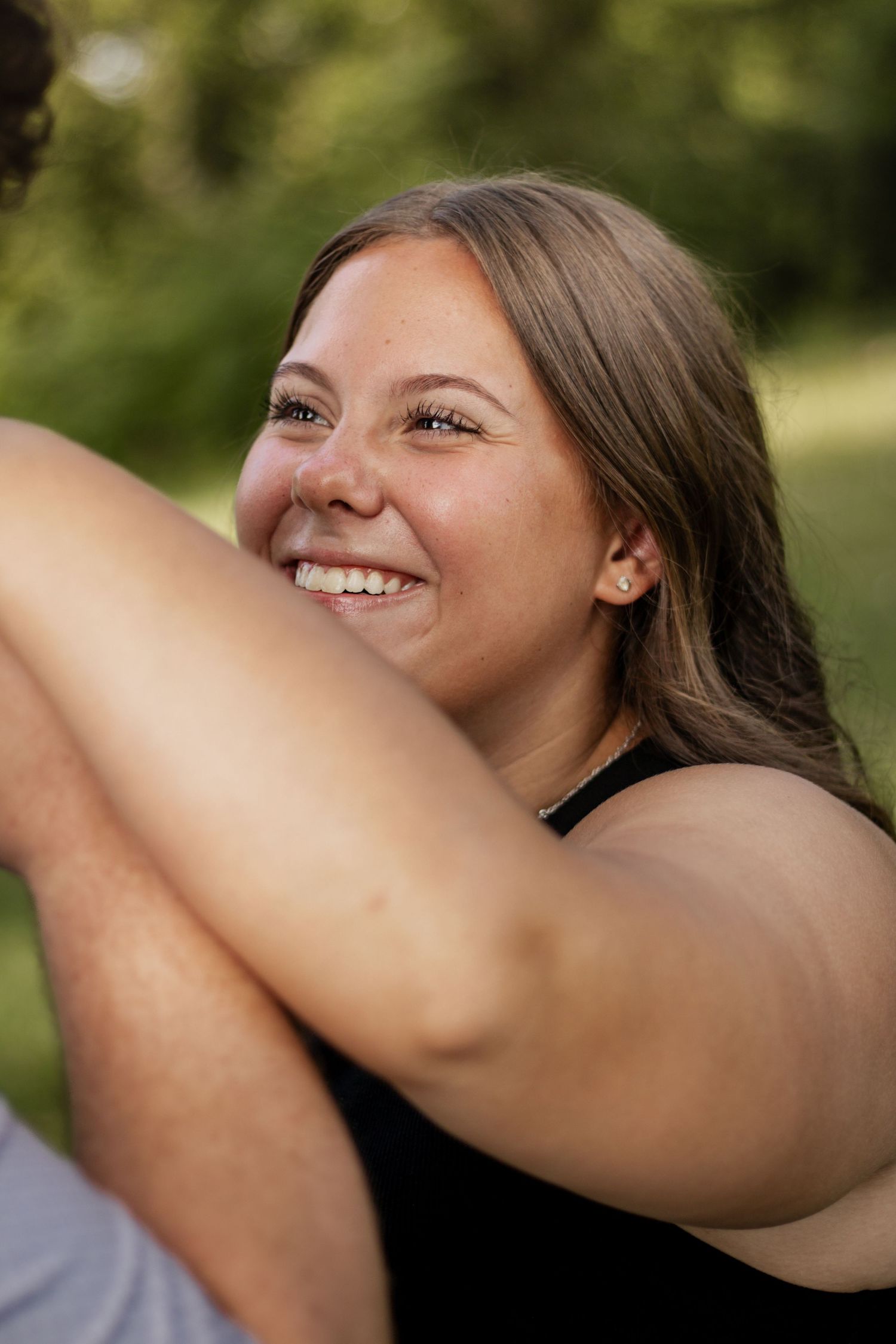 Smiling young woman with long brown hair sitting outdoors in a park, wearing a black top on a sunny day.