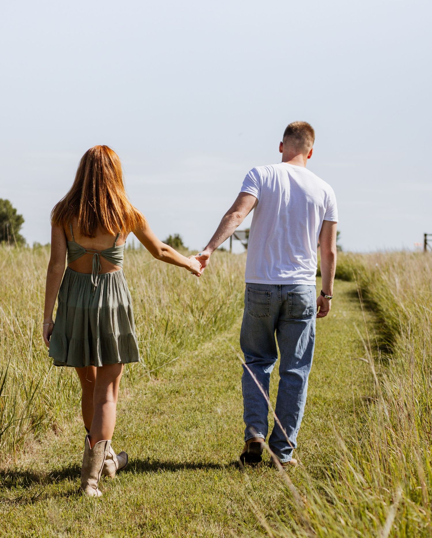Couple holding hands walking through a green grass field path on a sunny day, woman in green dress, man in jeans.