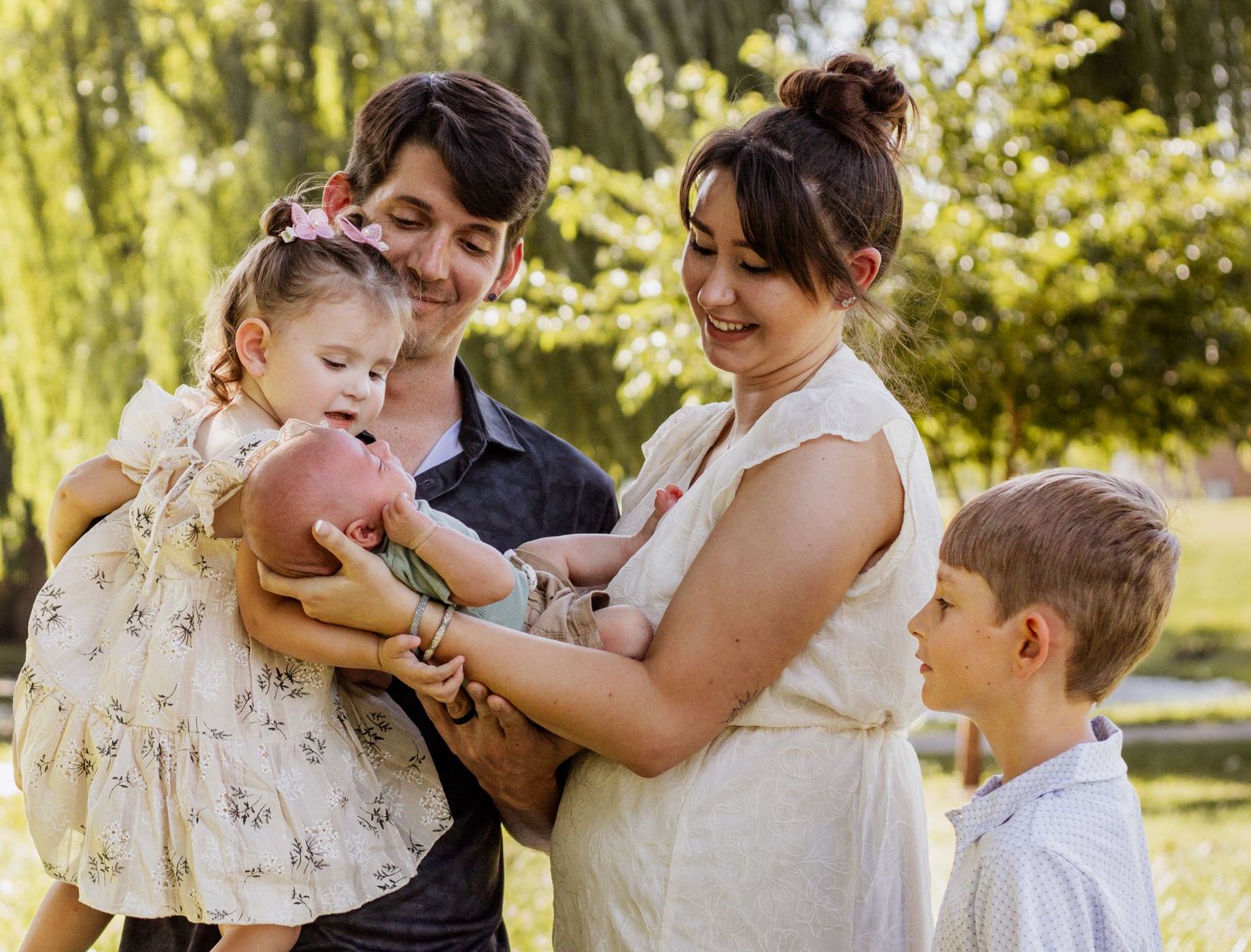 Family of four admiring newborn baby outdoors in sunny green park setting.