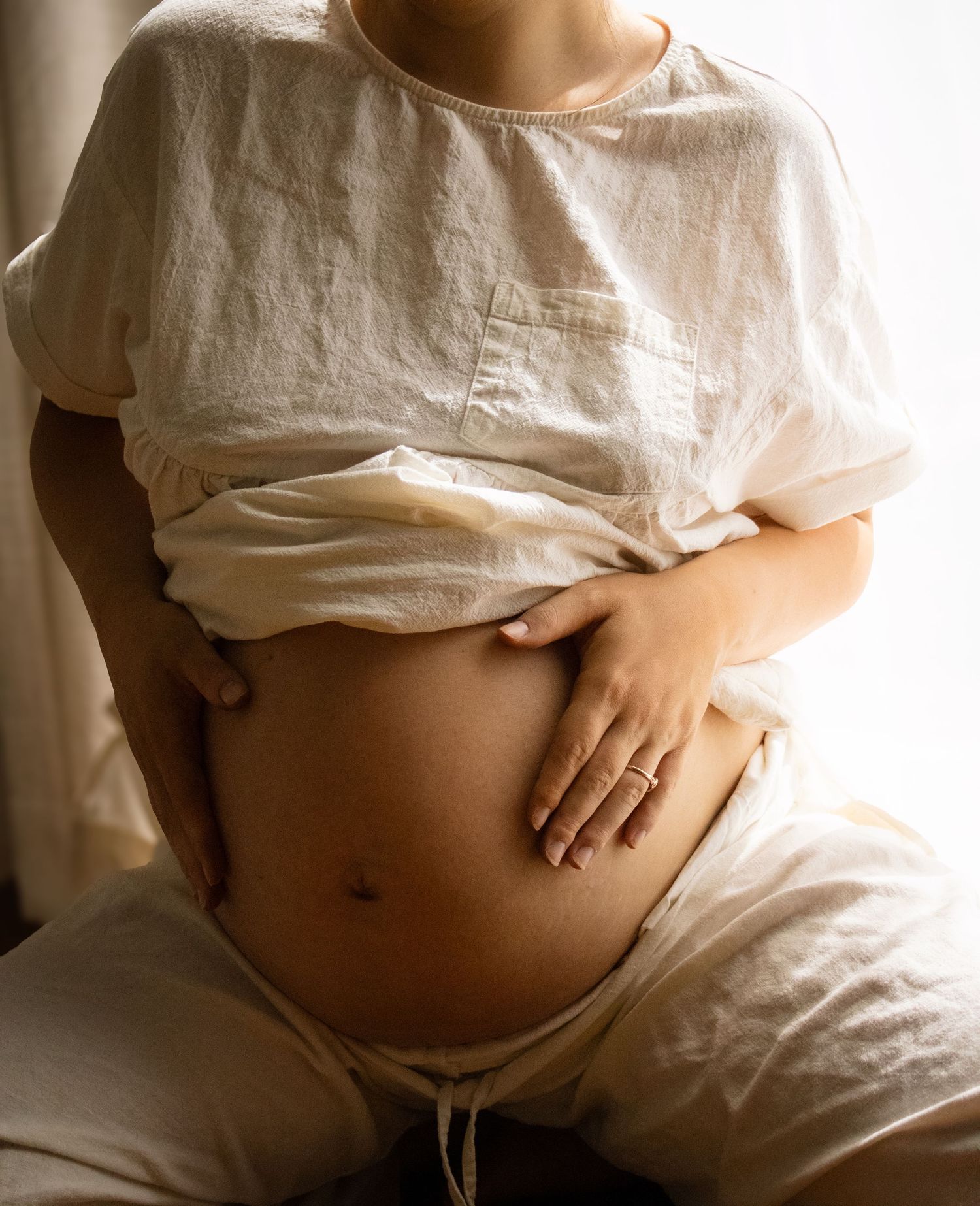 Close-up of pregnant woman in white top tenderly holding her bare round baby bump with both hands.