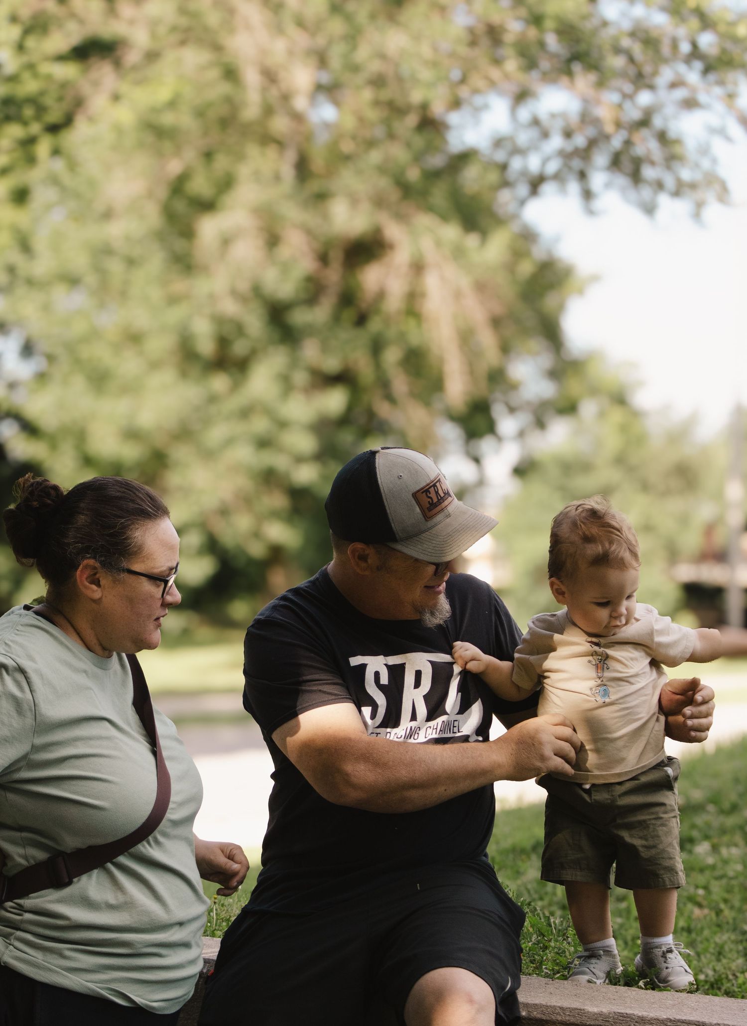 Two adults interacting with a standing toddler on a park bench surrounded by lush green trees.