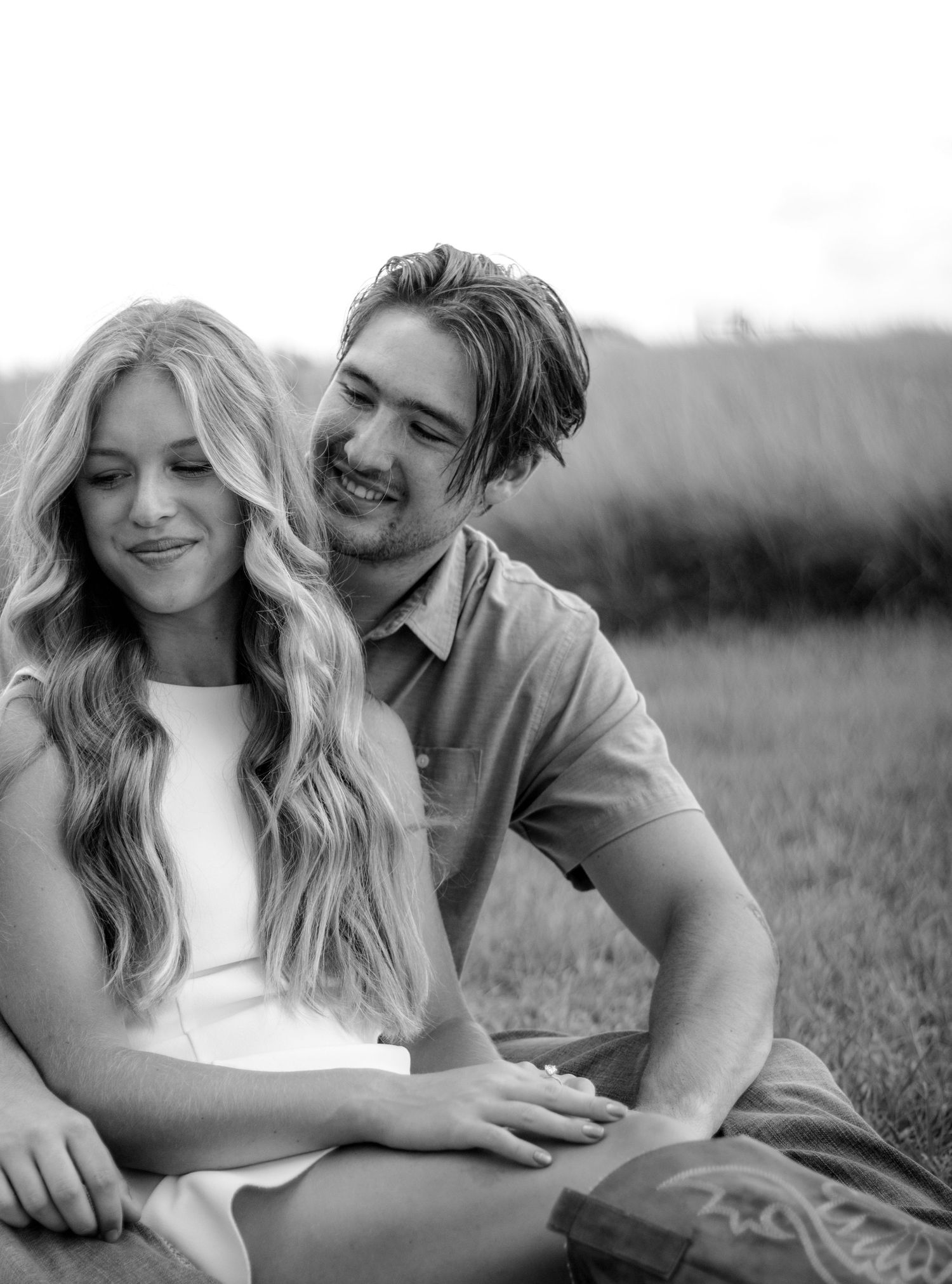 A happy couple sits together in a grassy field, the woman leaning into the man in this romantic black and white photo.