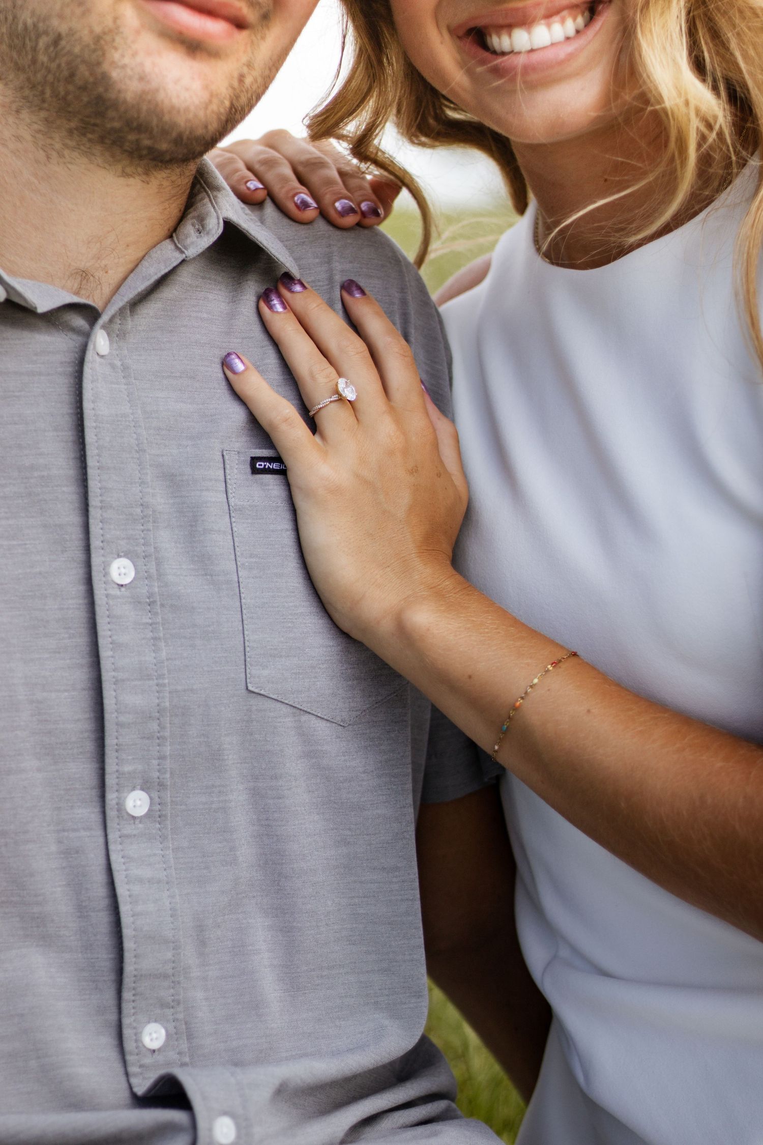 Smiling woman with purple nails and diamond engagement ring rests hand on man's gray Patagonia button-up shirt.