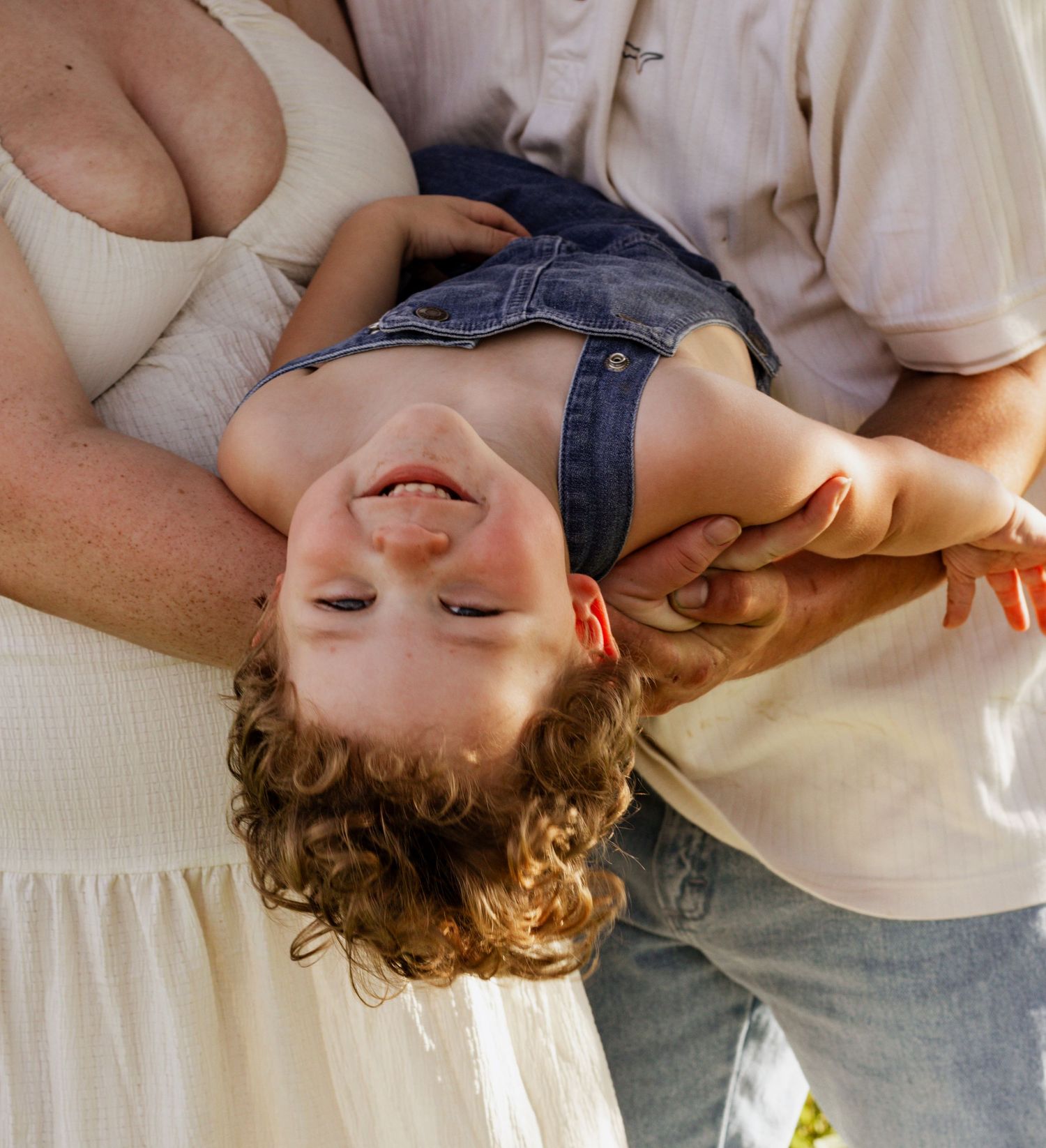Parents playfully holding their curly-haired toddler upside down, laughing together outdoors in warm sunlight.