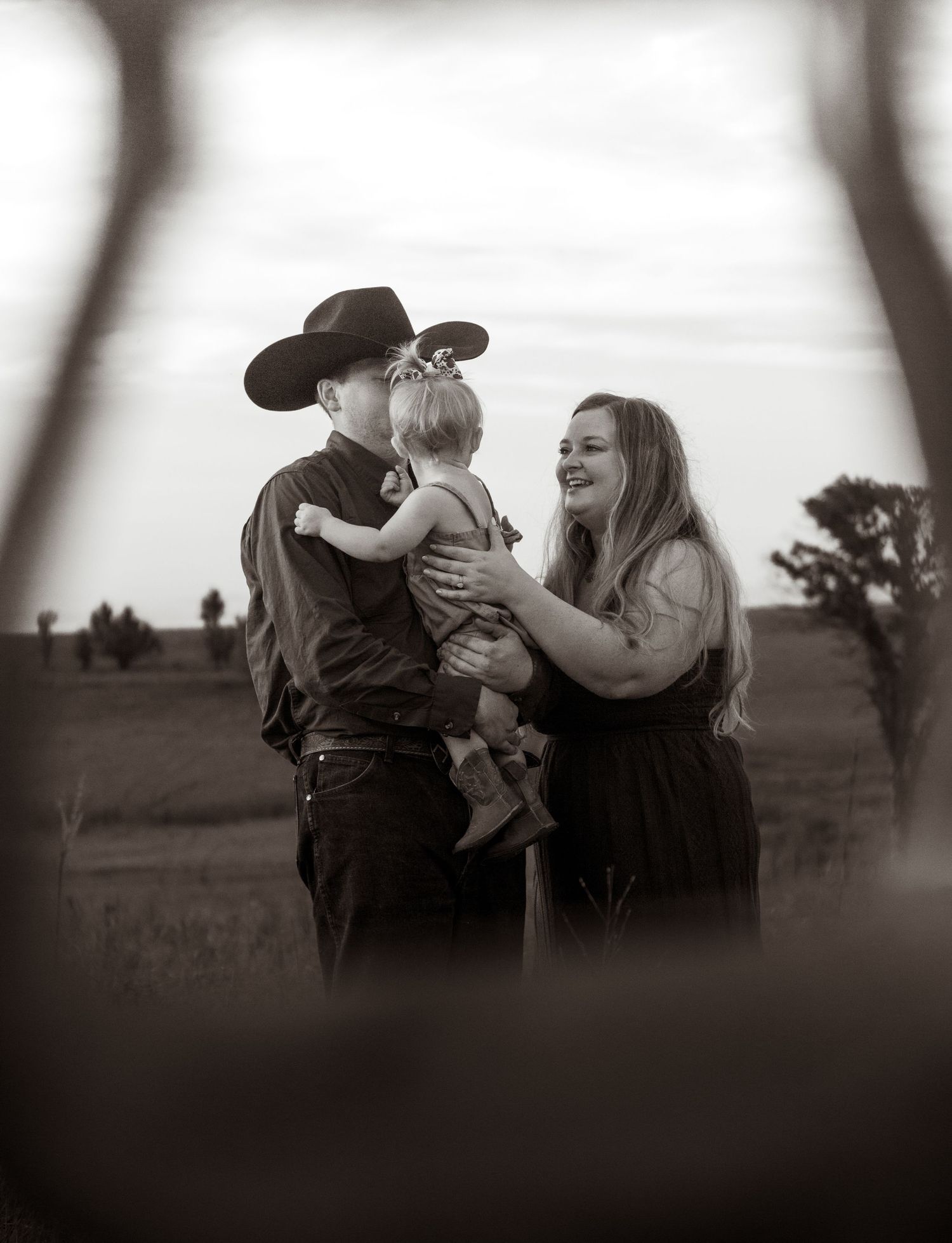 Black and white photo of a man in a cowboy hat kissing a toddler while a smiling woman stands beside them outdoors.