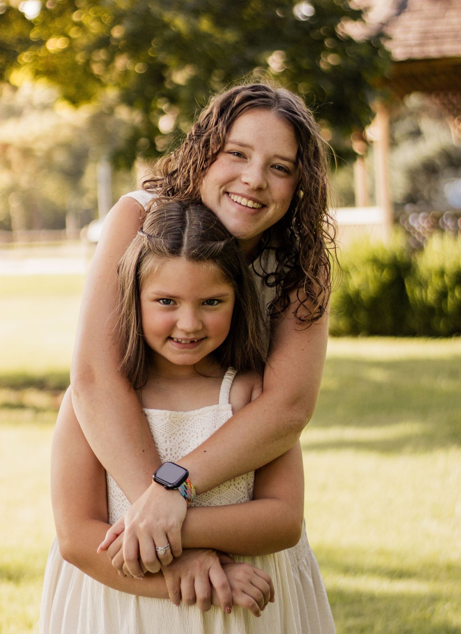 Two smiling girls in white dresses embrace outdoors in a sunny green park setting.