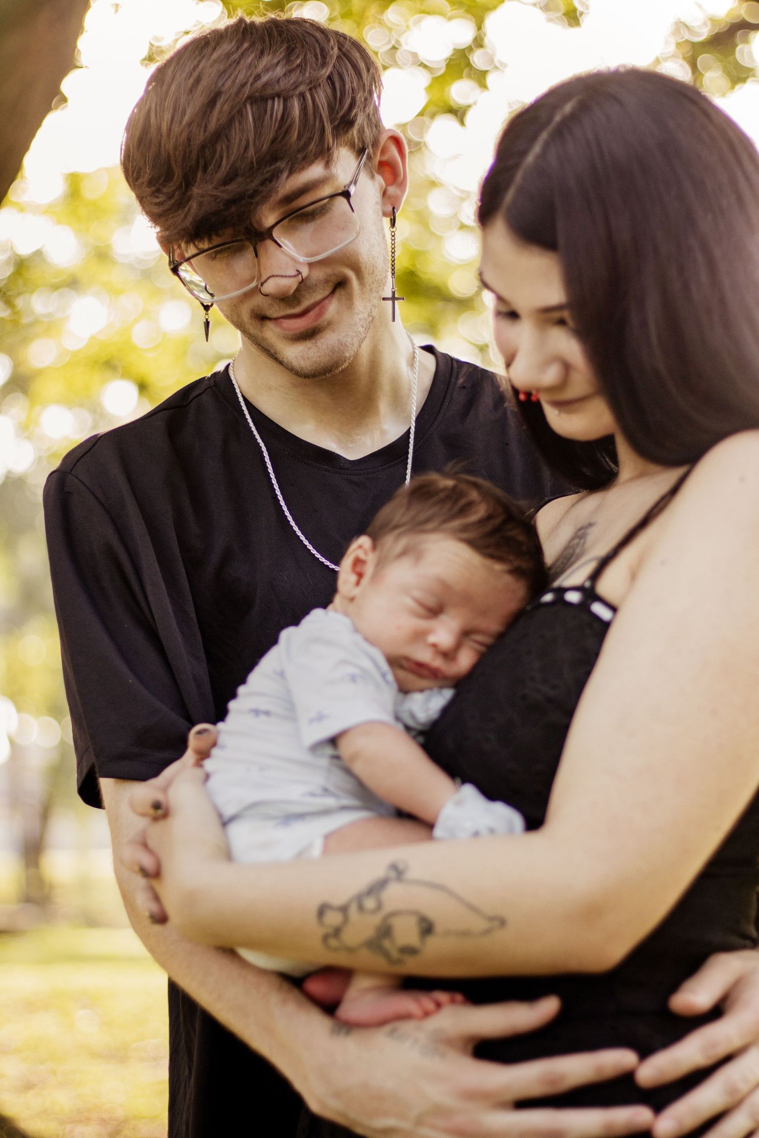 Young couple with glasses and tattoos lovingly holding their sleeping newborn baby outdoors in a sunny park.