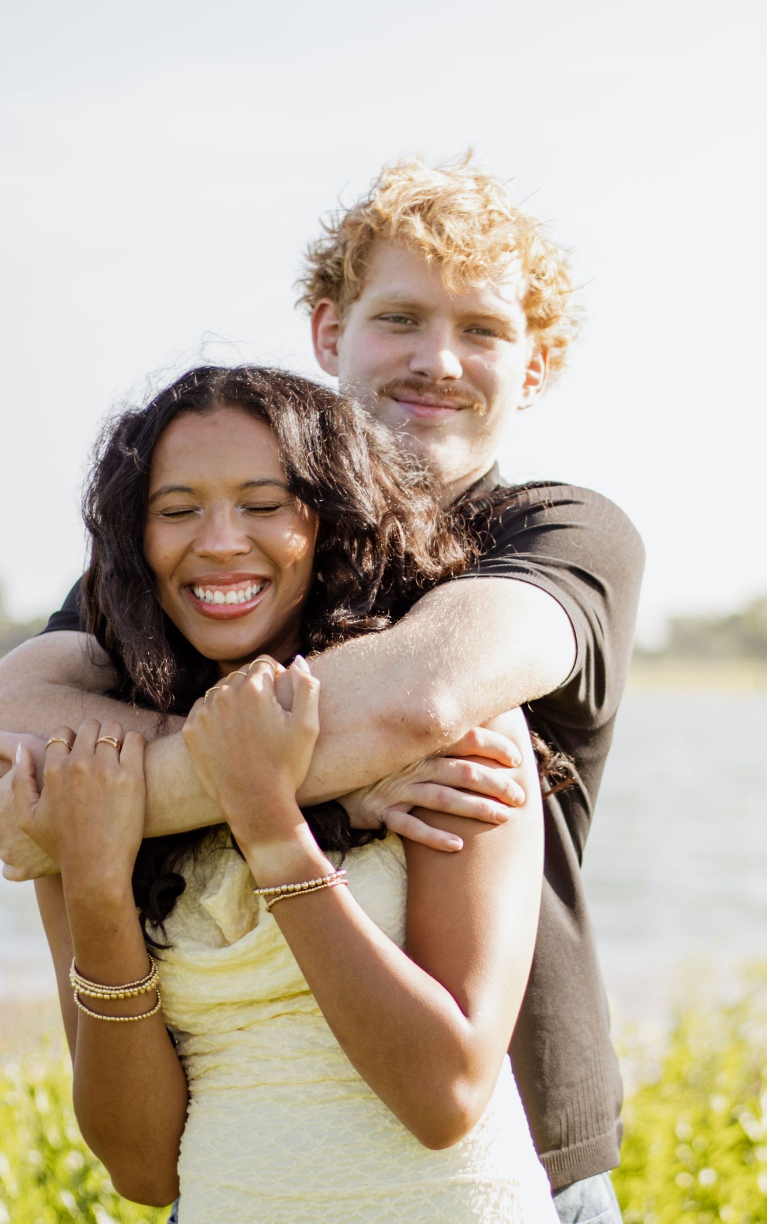 A happy interracial couple embracing outdoors, smiling man with curly red hair hugging a laughing woman in a yellow dress.