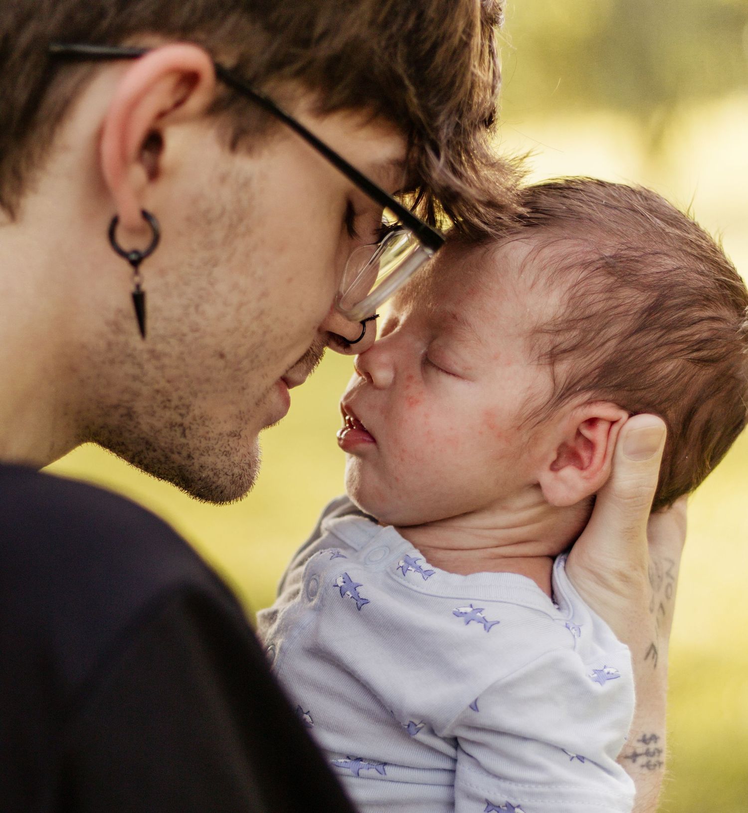 A young tattooed man with glasses and hoop earring tenderly touches foreheads with a sleeping newborn baby outdoors.