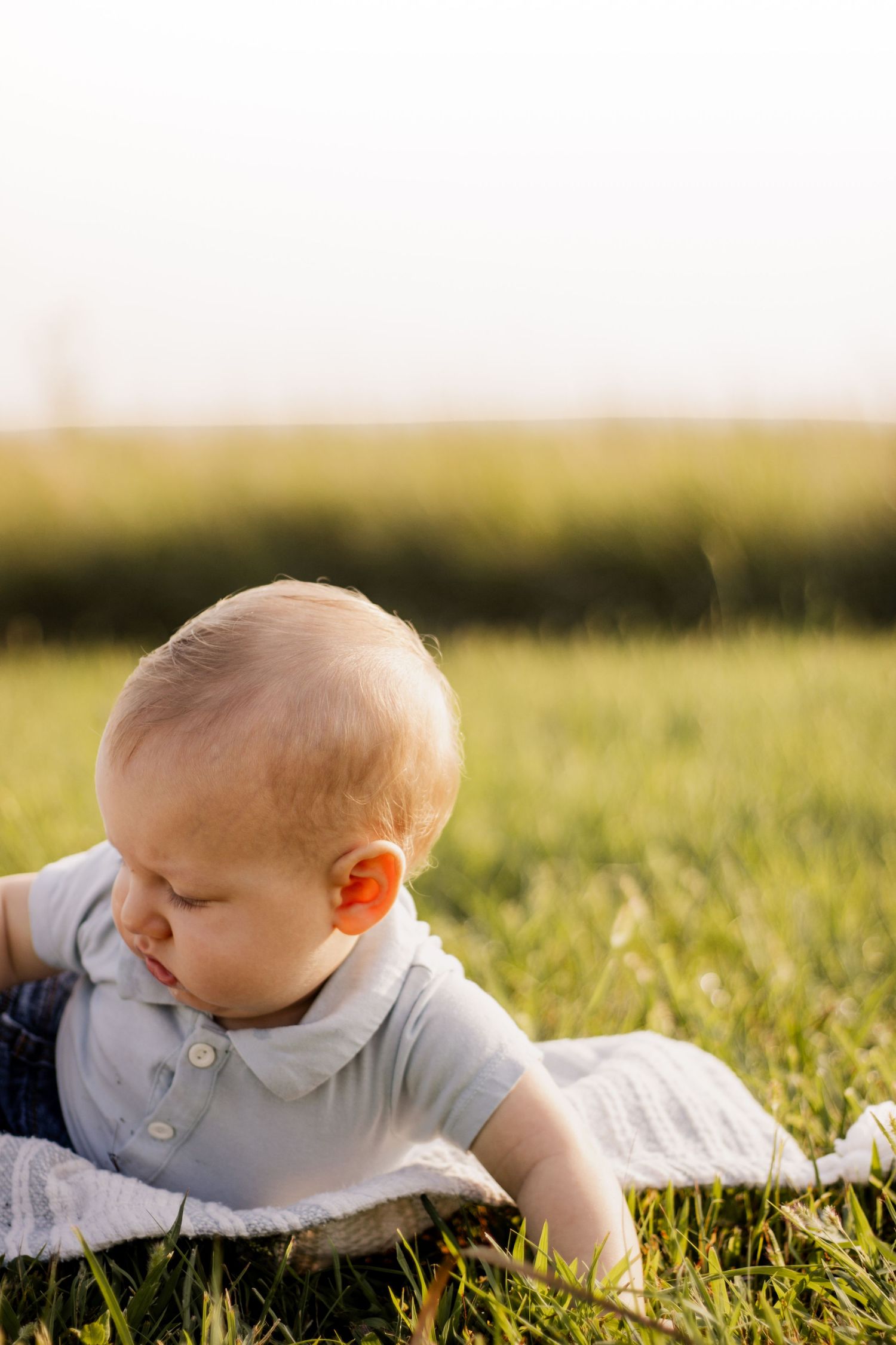A baby boy lies on a white blanket in a sunny green grass field, looking down curiously.