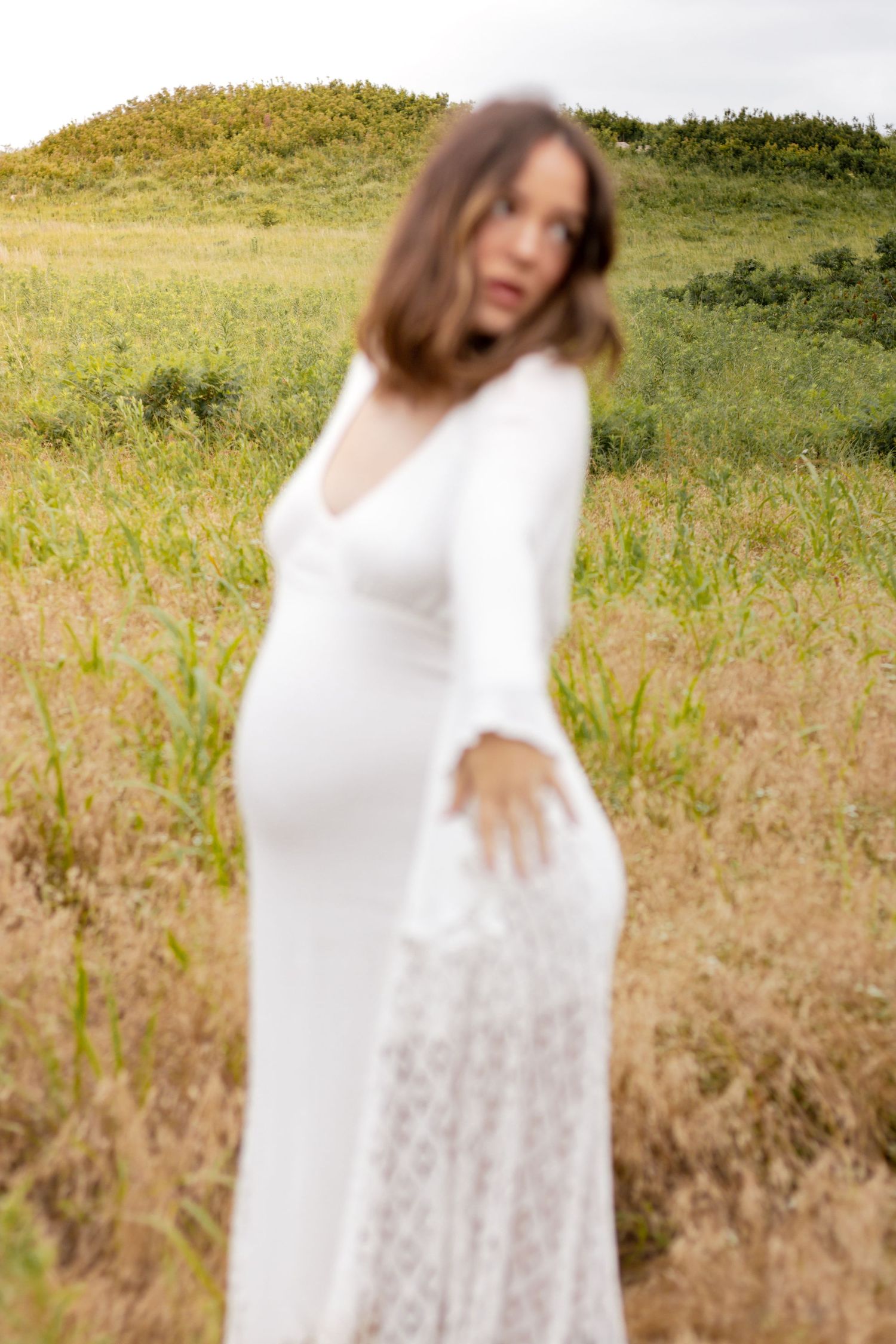Blurred foreground of pregnant woman in white dress standing in a golden grass field with green hills.