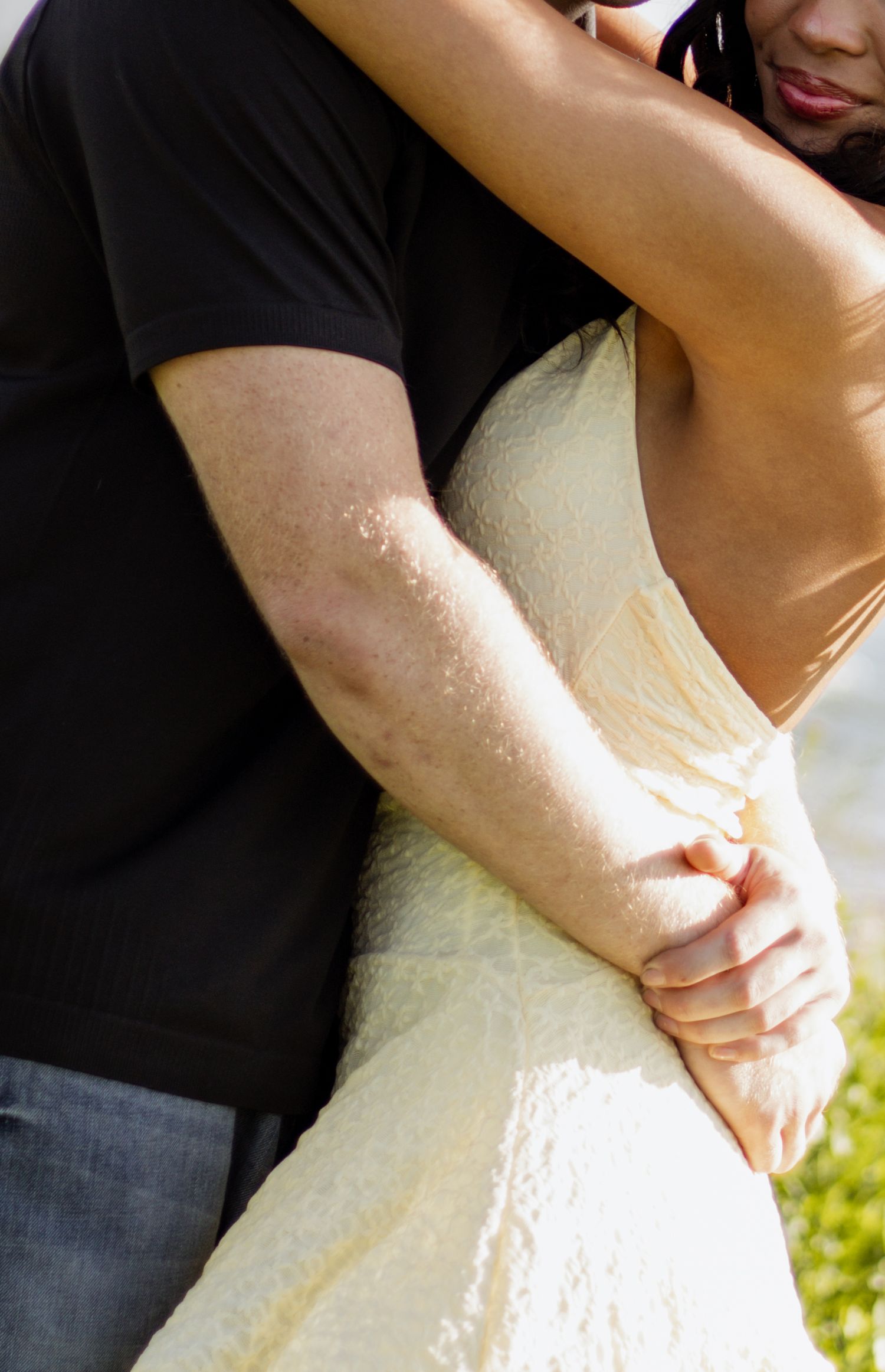 A couple embraces tenderly, the man in a black shirt holding a woman in a white lace dress outdoors.