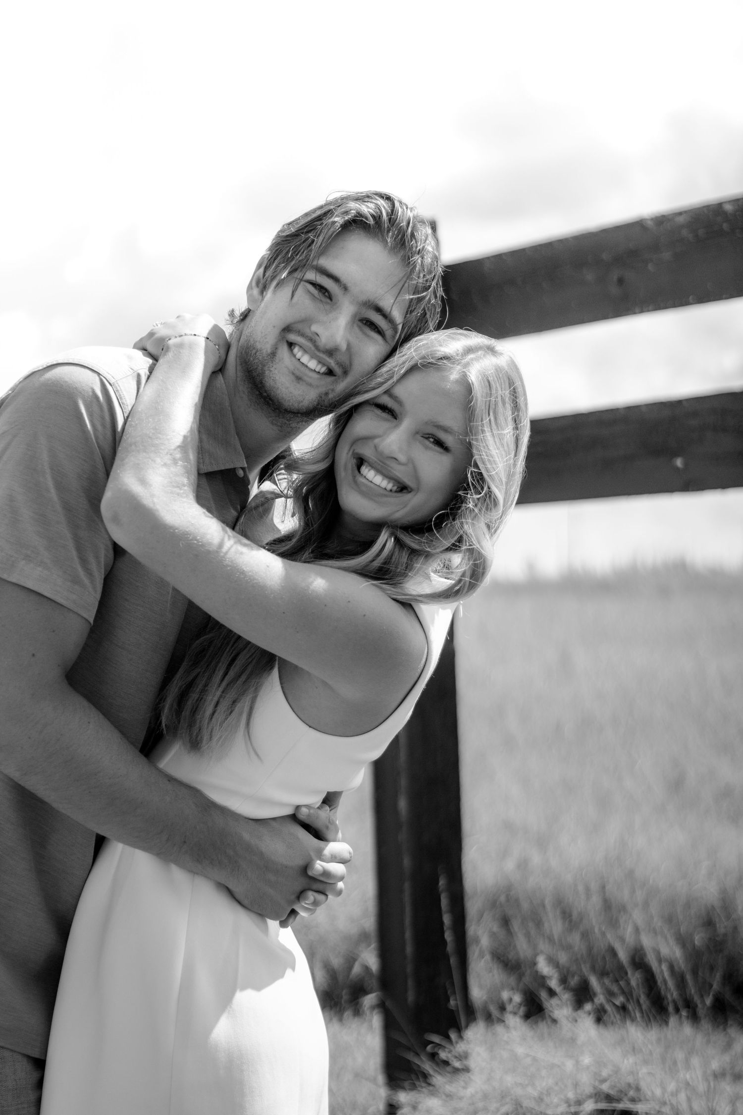 Smiling couple embracing near a wooden fence in a black and white outdoor engagement photo.