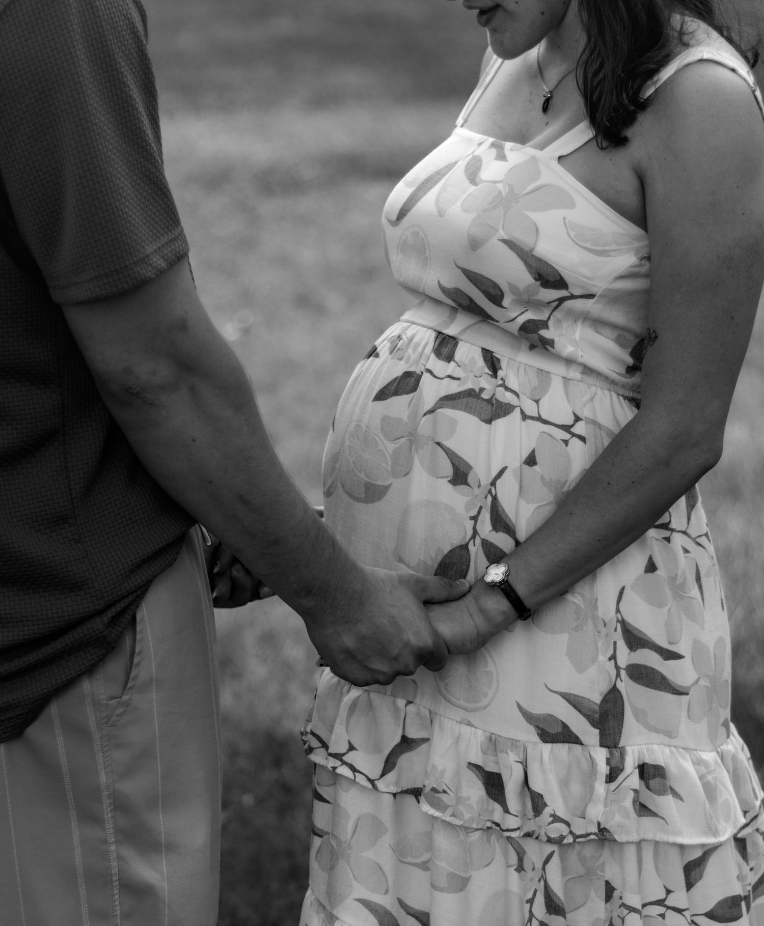 Black and white photo of a pregnant woman in floral dress holding hands with her partner outdoors.