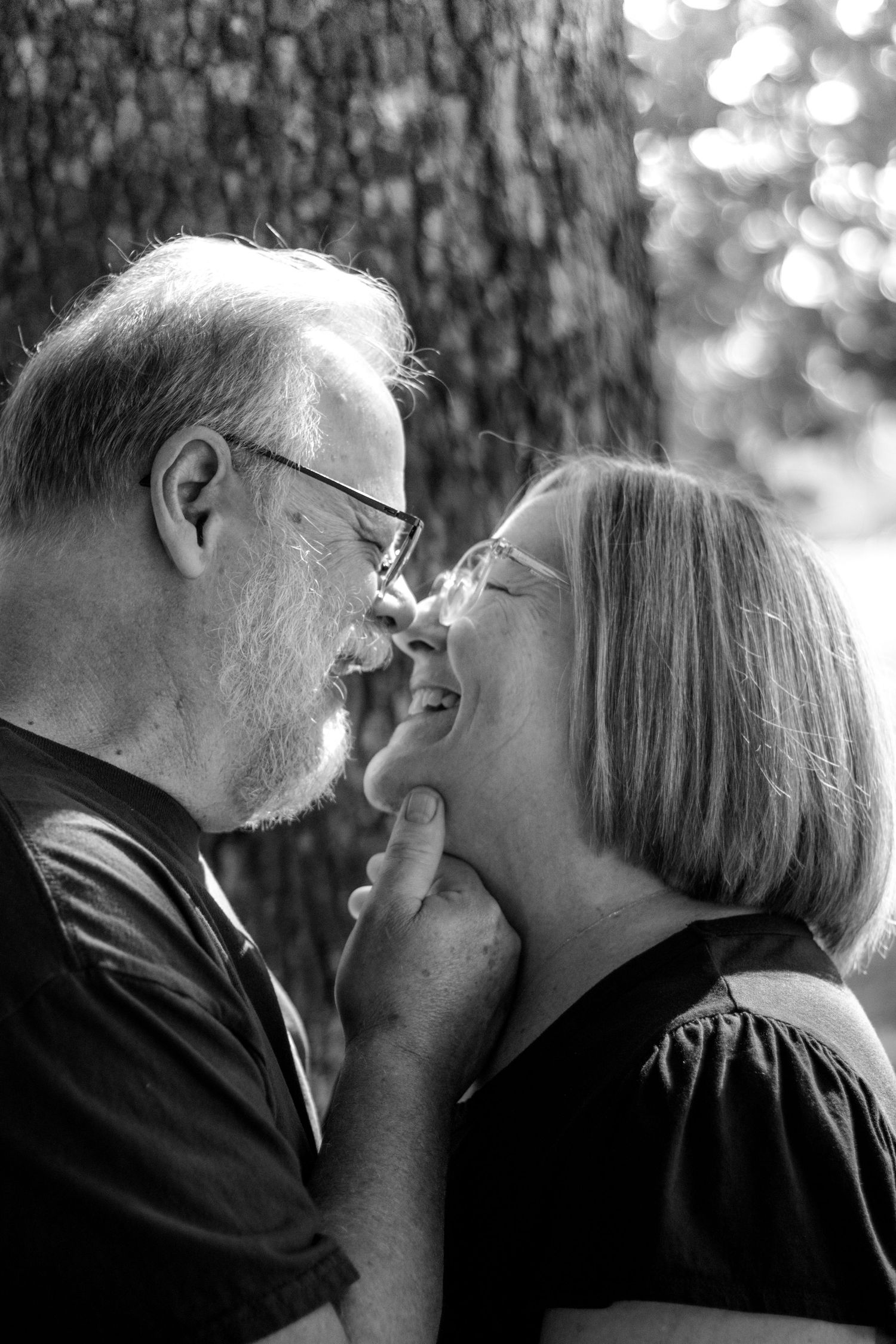 Black and white photo of a mature couple sharing a tender moment, smiling and nearly kissing outdoors near a tree.