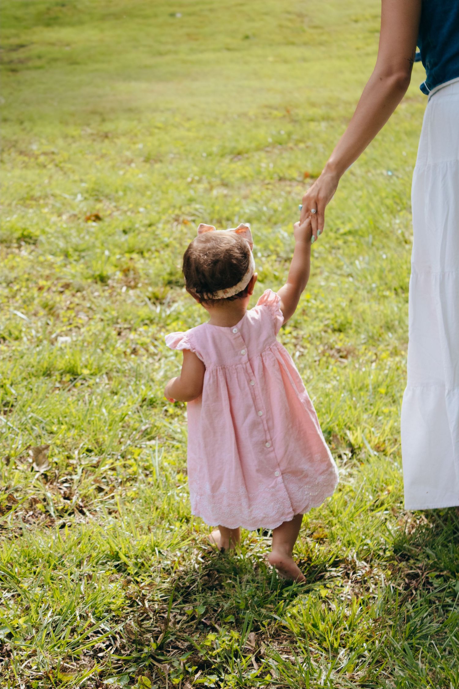 A toddler girl in a pink dress holds her mother's hand while walking barefoot on green grass.