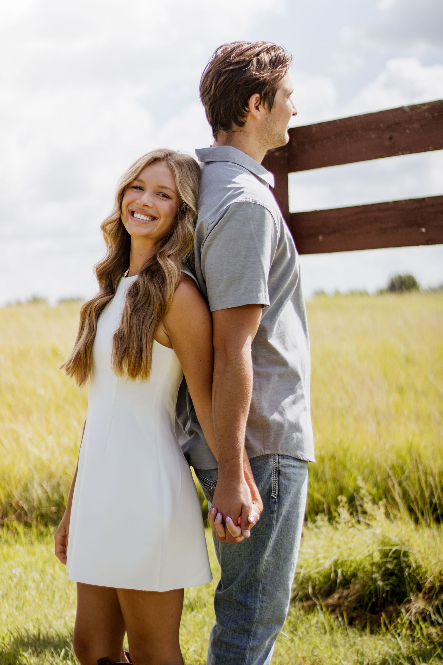 A smiling couple holds hands in a sunny field near a wooden fence, the woman in a white dress and man in gray shirt.