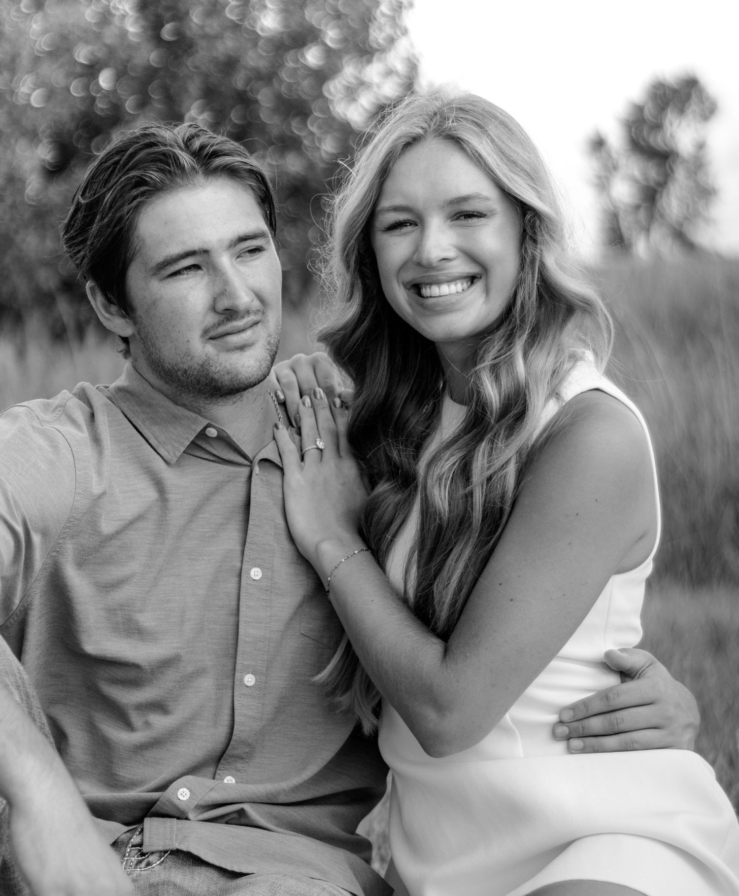Black and white photo of a smiling couple posing outdoors, woman showing engagement ring while leaning on mans shoulder.