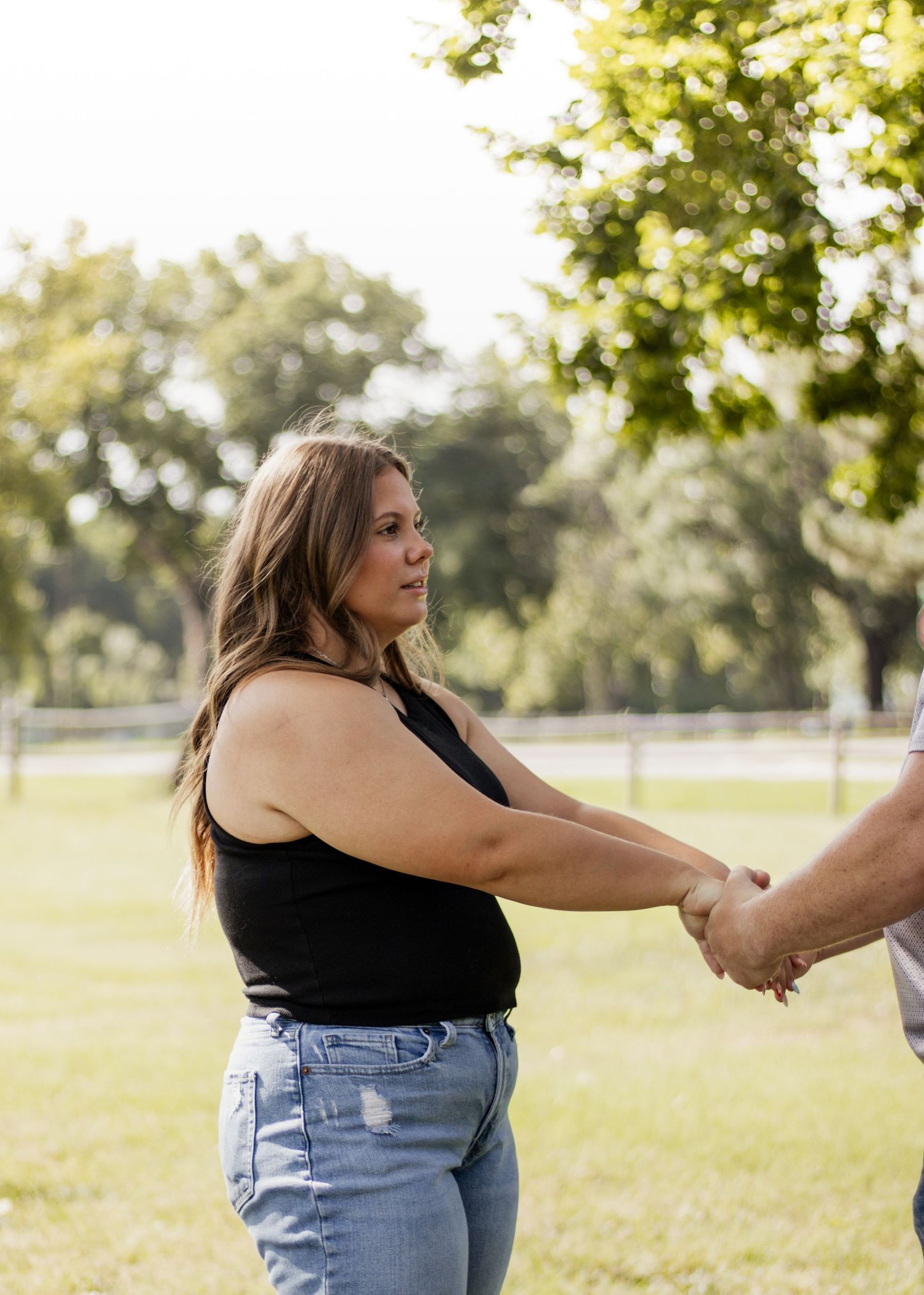 A woman in a black tank top and jeans holds hands with someone outdoors in a sunny green park.