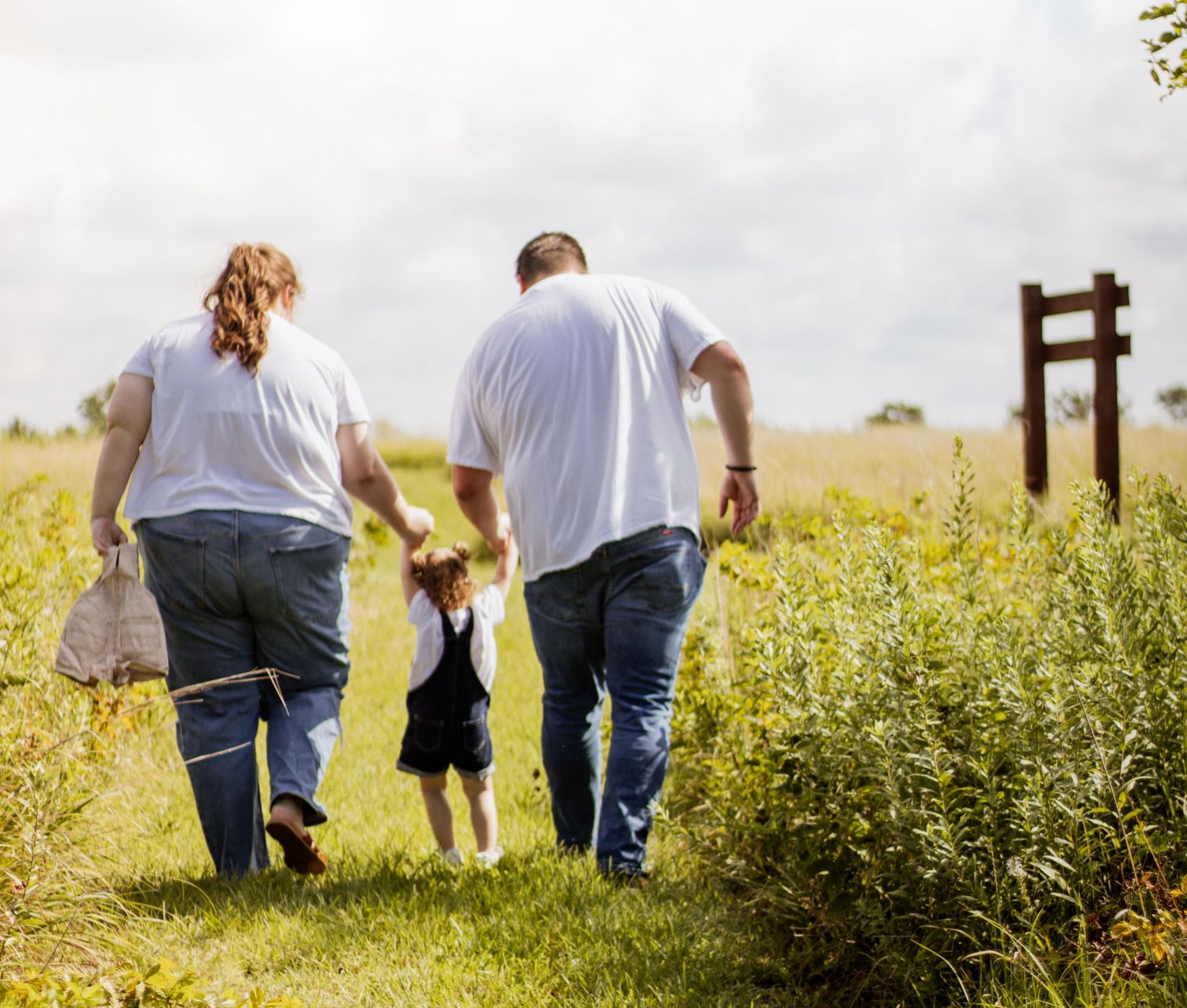 Family of three walking hand-in-hand through a lush green field on a cloudy day.