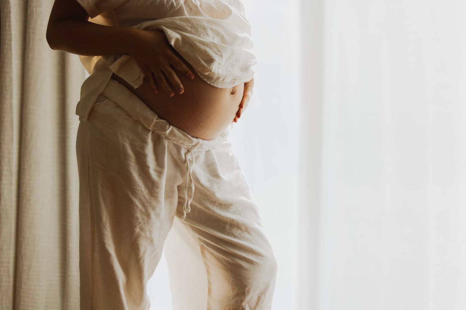 Pregnant woman in white linen pants and top holds baby bump near sunlit window with sheer curtains.