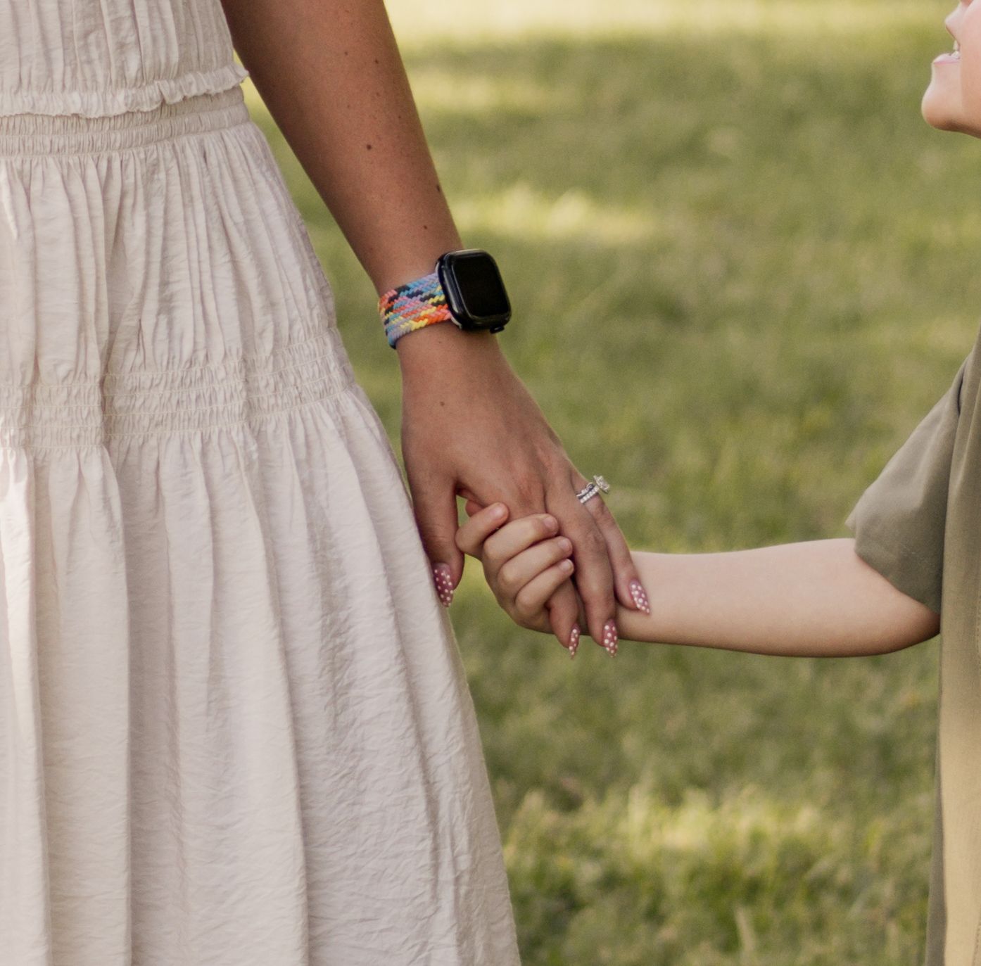 Adult and child holding hands outdoors, adult wearing a smartwatch and white dress, child in olive green shirt.