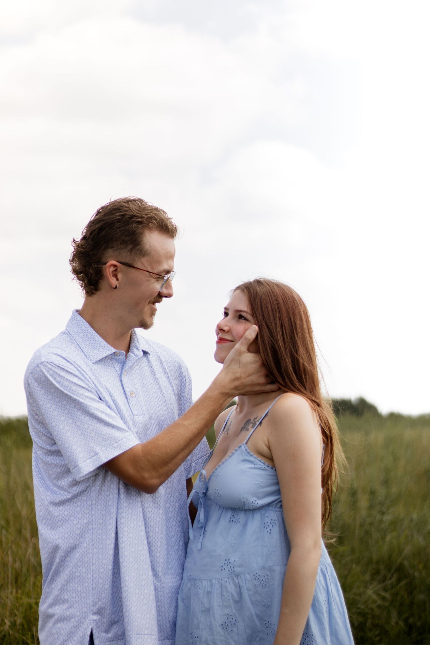 A young couple shares a tender moment in a green field, the man gently touching the woman's face as they smile at each other.