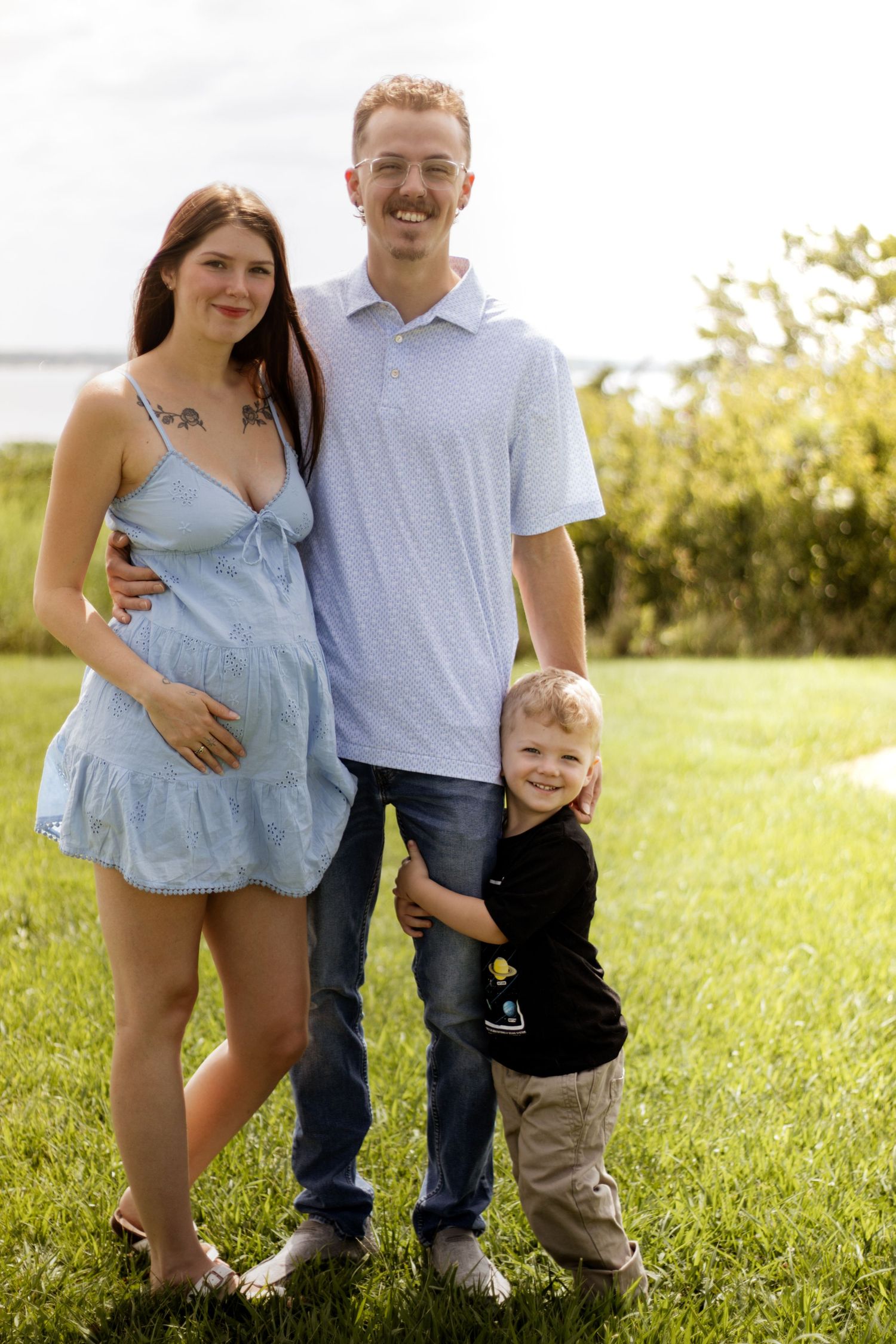 Smiling pregnant couple with young boy posing outdoors on green grass near water on a sunny day.