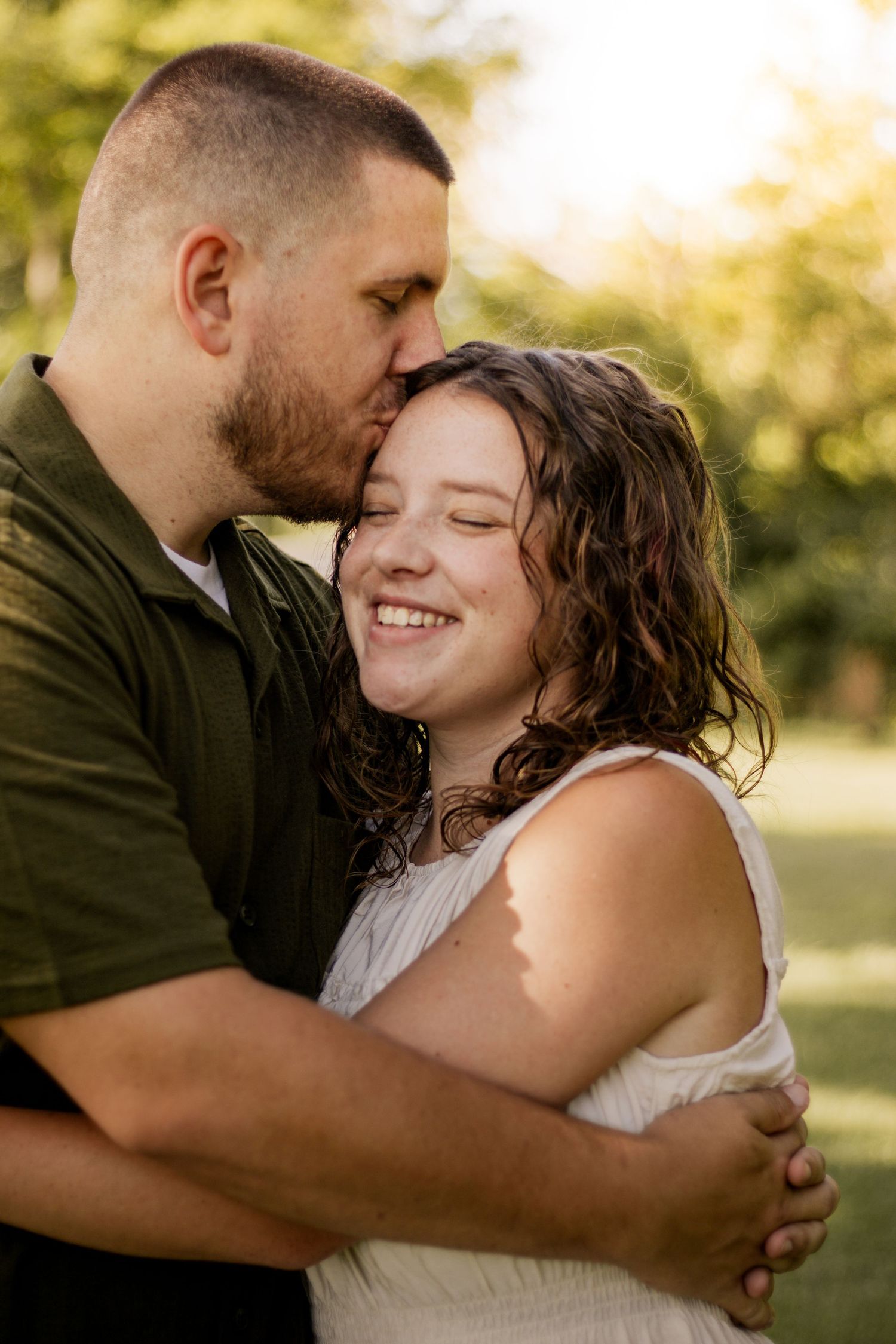 A couple embraces outdoors as a man kisses a smiling woman on the cheek in golden sunlight.