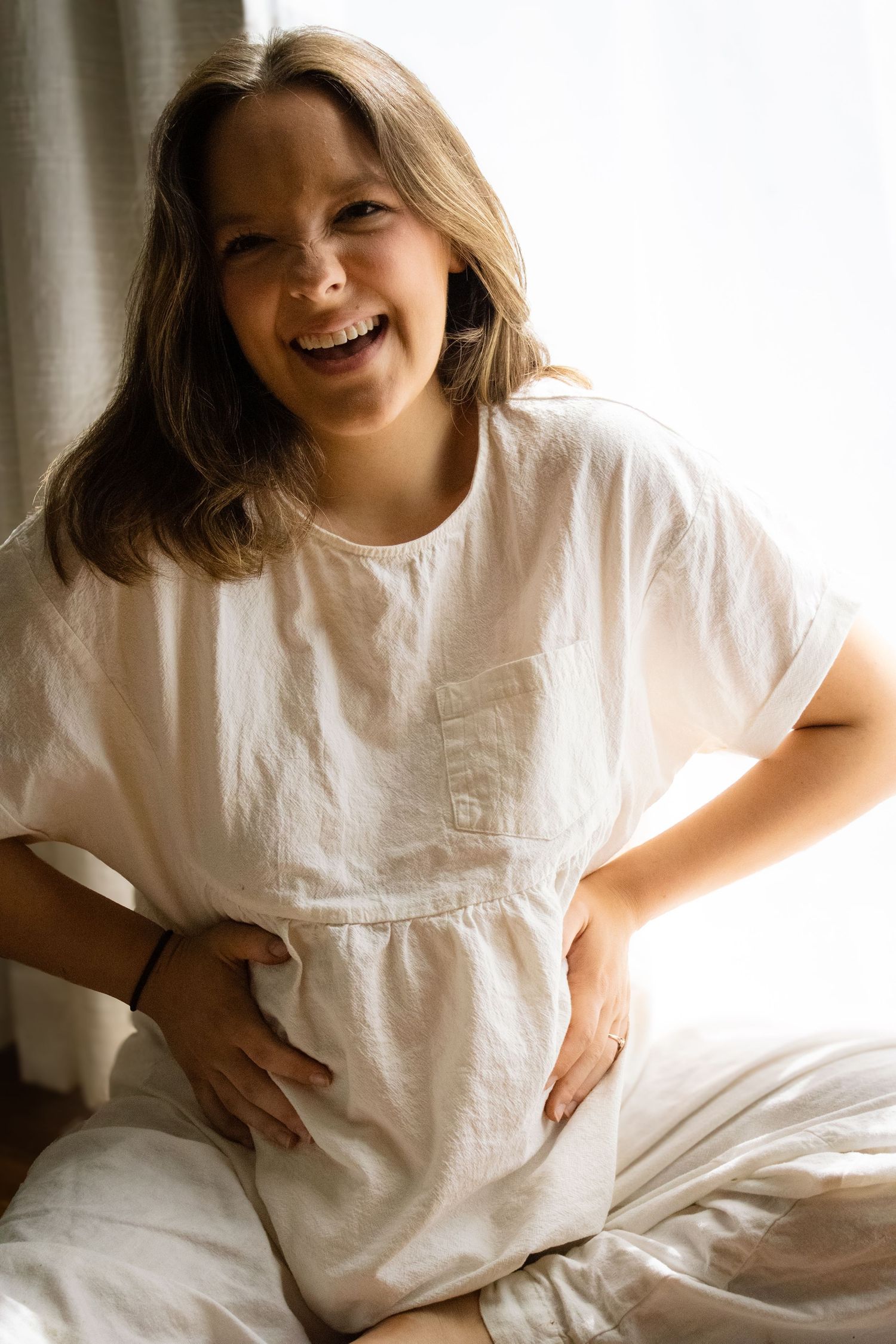 Happy laughing pregnant woman in white linen dress sitting on bed holding her baby bump.