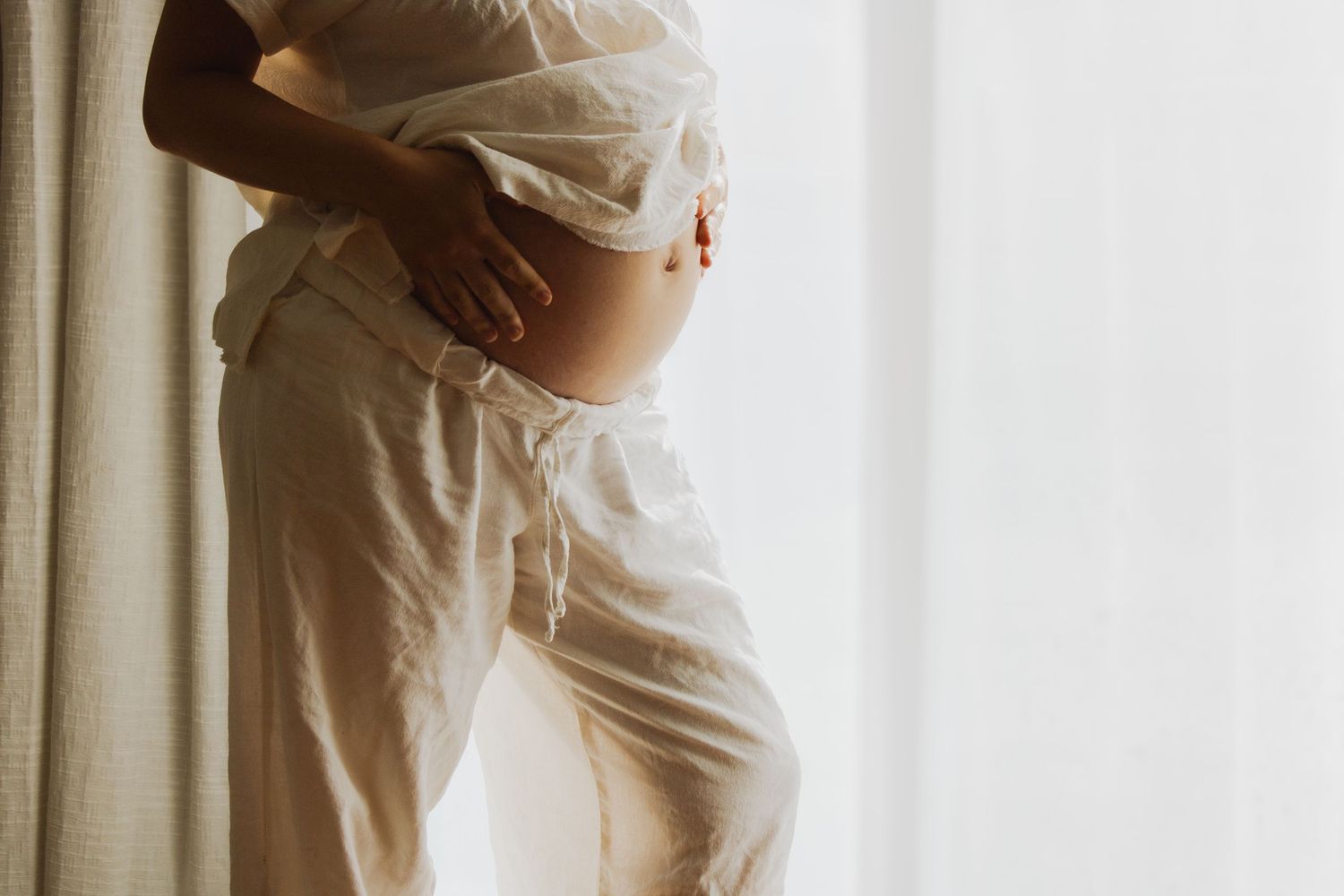 Pregnant woman in white linen ensemble cradles bump standing beside natural light curtained window.
