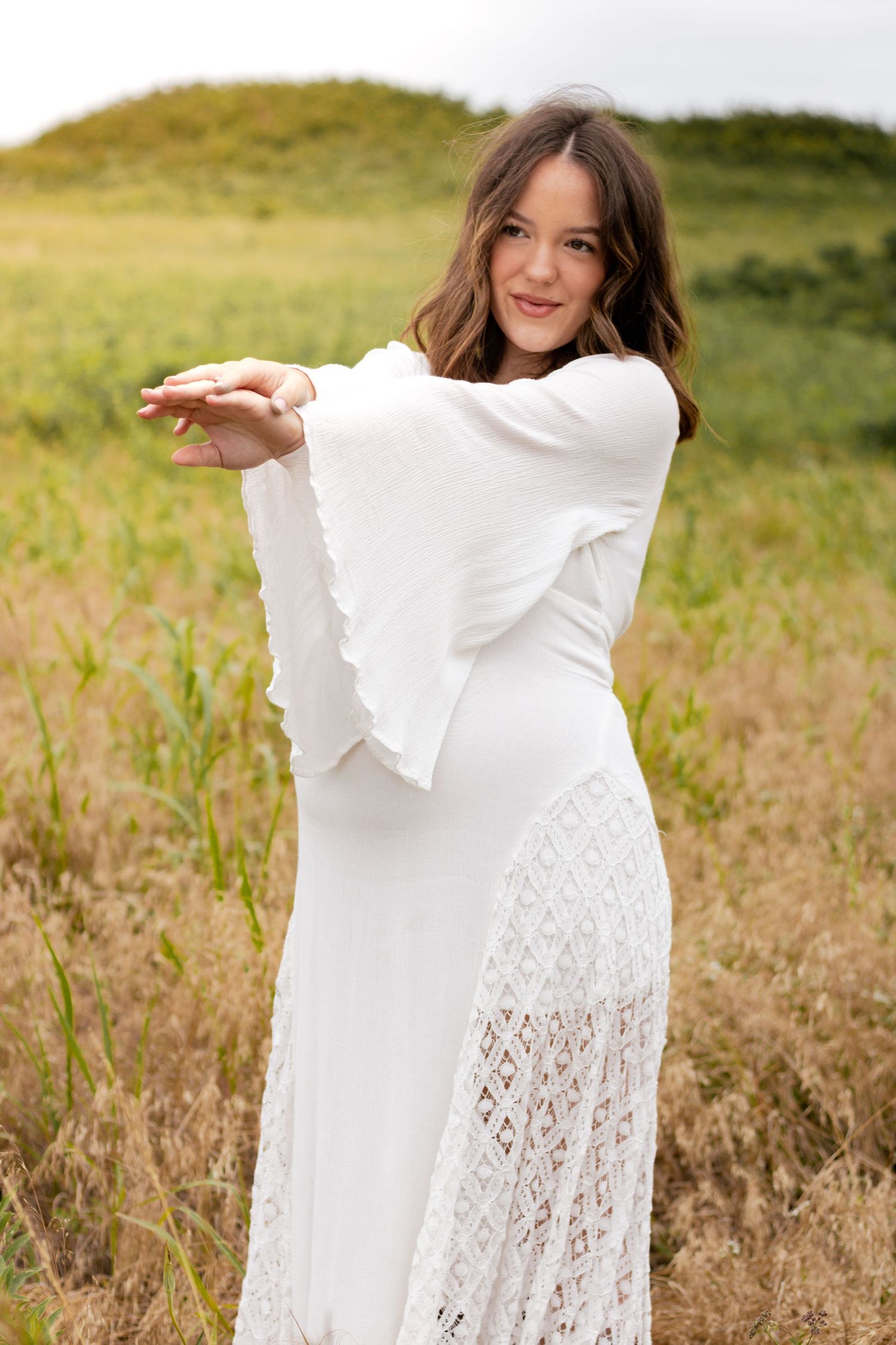 Smiling woman in white lace dress stretching arms forward in grassy field.