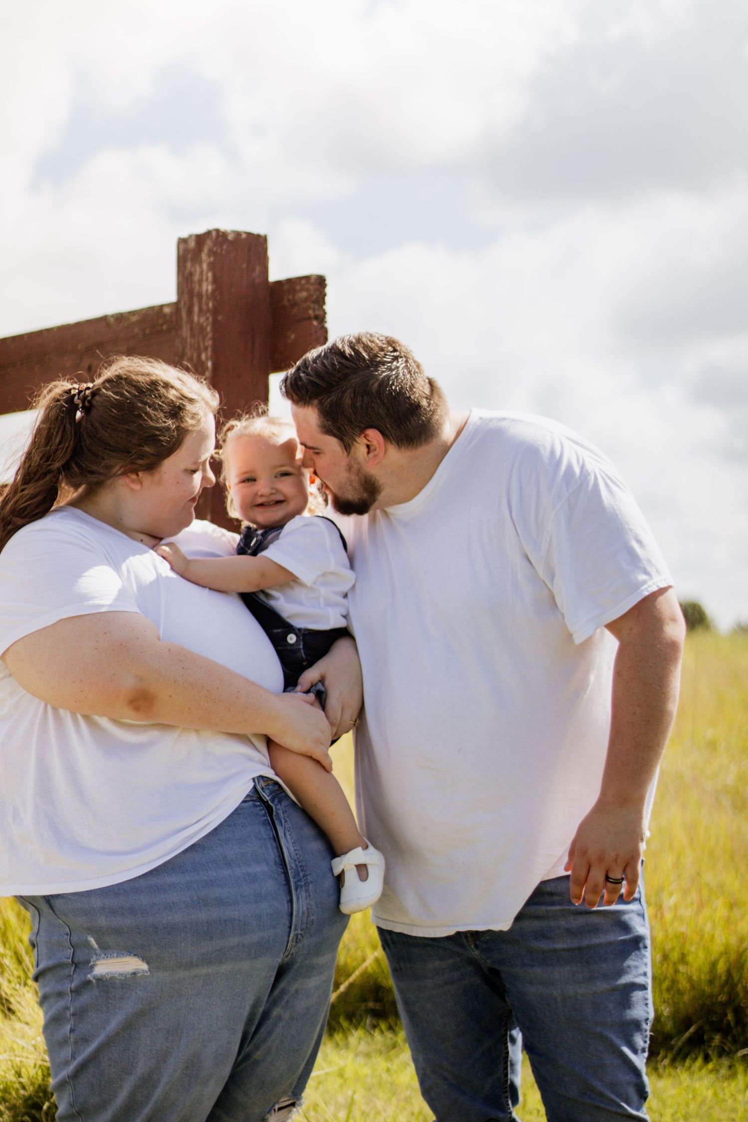 A young family with a baby outdoors near a wooden fence, parents in white tees and jeans, dad kissing the smiling infant.