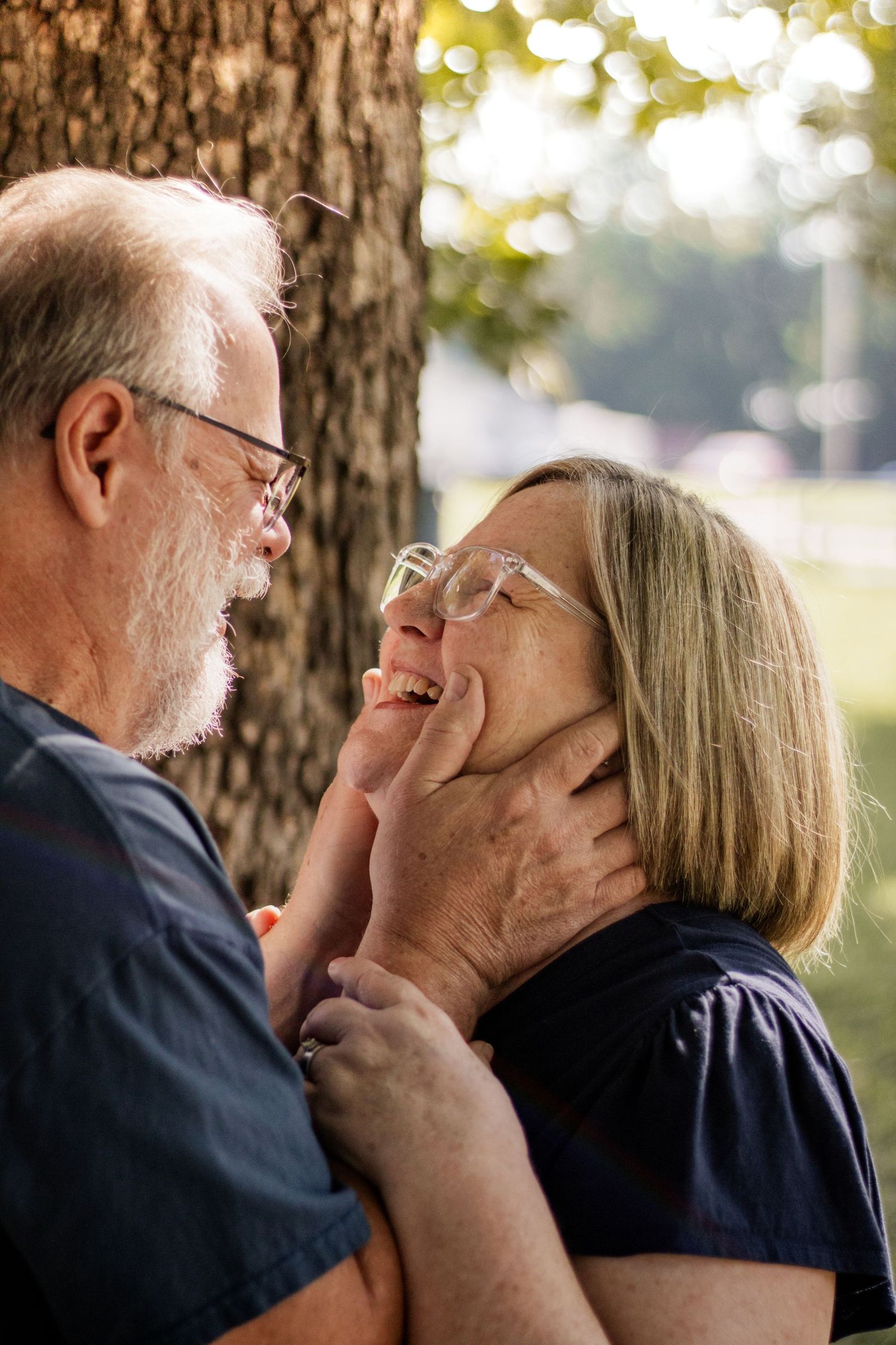 Elderly couple sharing a tender moment outdoors, man gently cupping womans face as they smile at each other near a tree.
