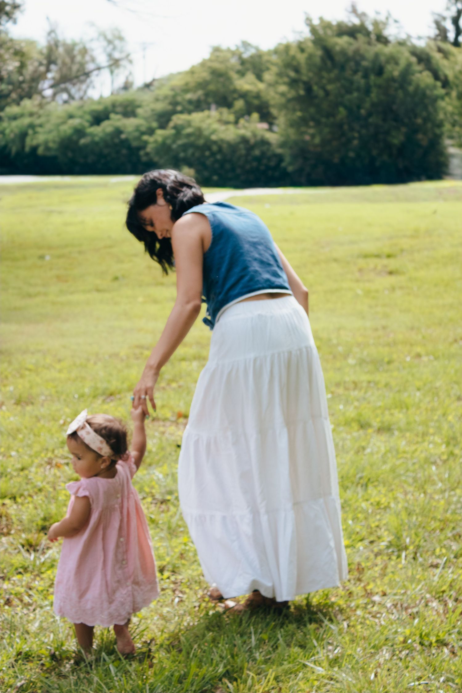 Mother in blue top and white skirt helping toddler girl walk on green grass in park.