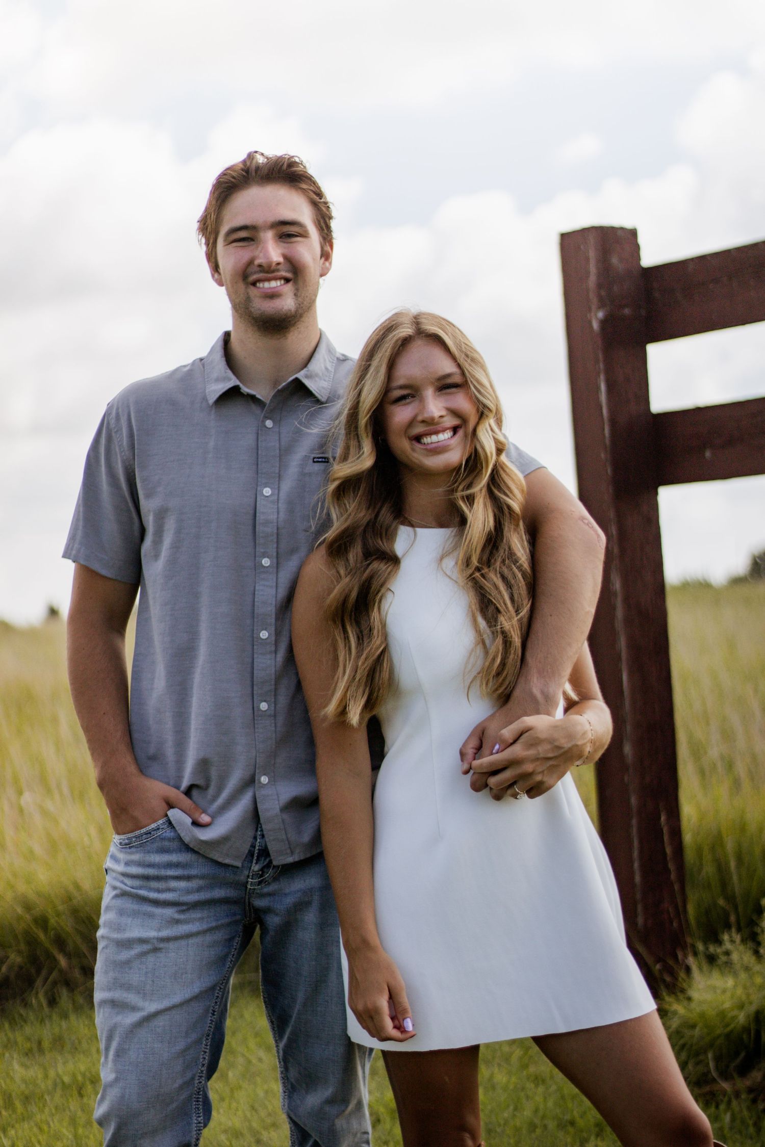 A smiling young couple poses outdoors in a green field, man in gray shirt and jeans, woman in white mini dress.