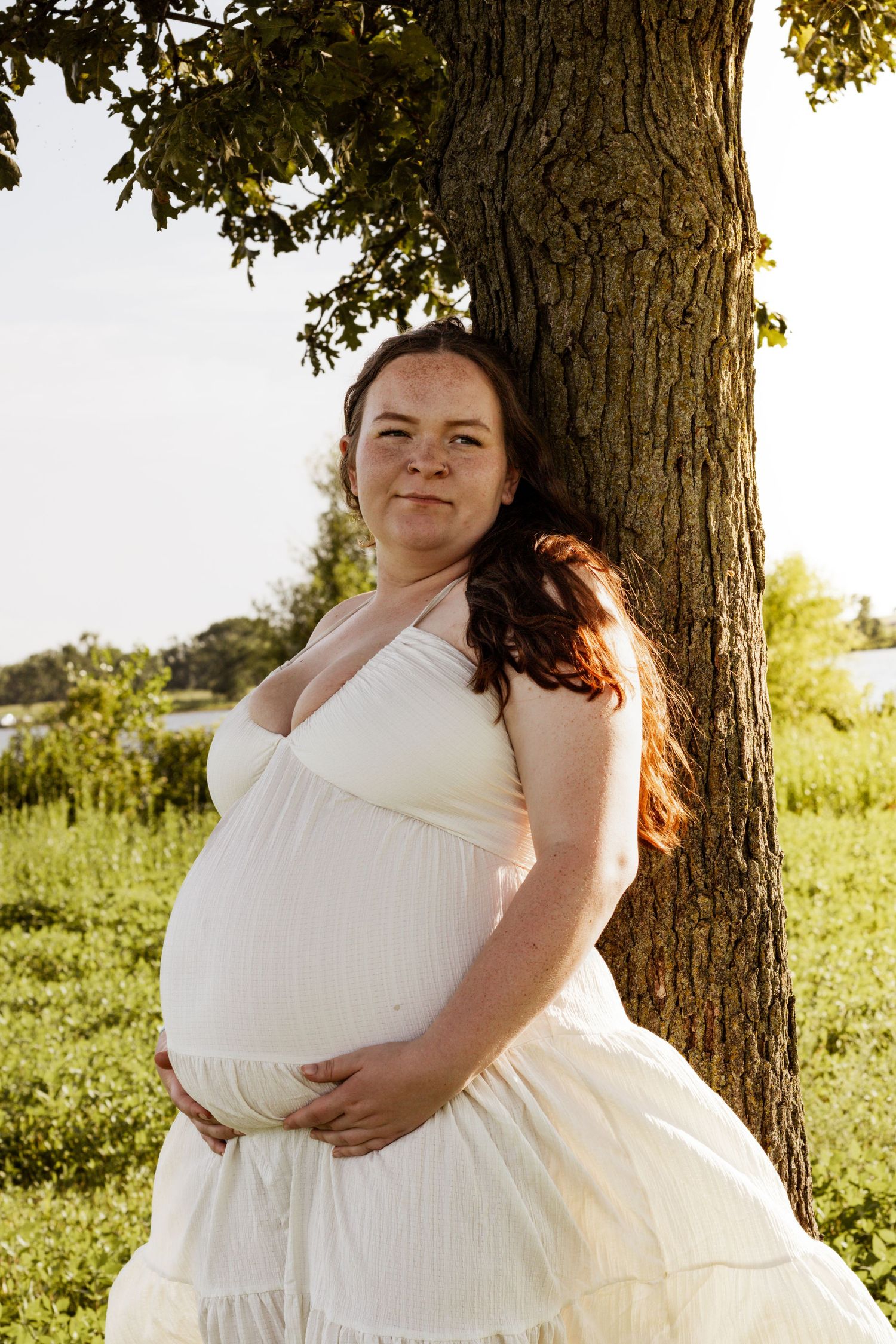 Pregnant woman in white dress leaning against a tree outdoors, cradling her baby bump in golden sunlight.