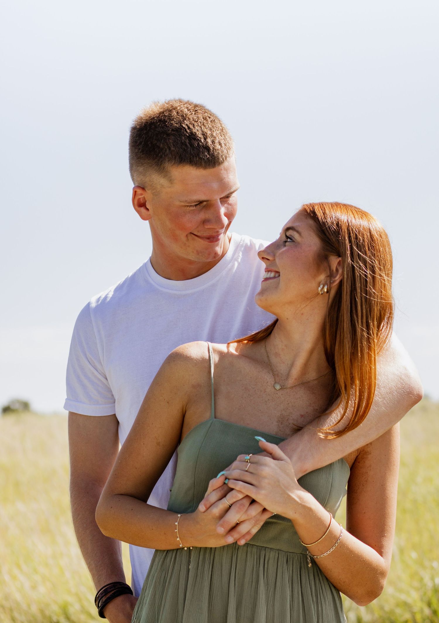 A happy young couple embraces in a sunny green field, the woman in a sage dress smiling up at the man in a white tee.