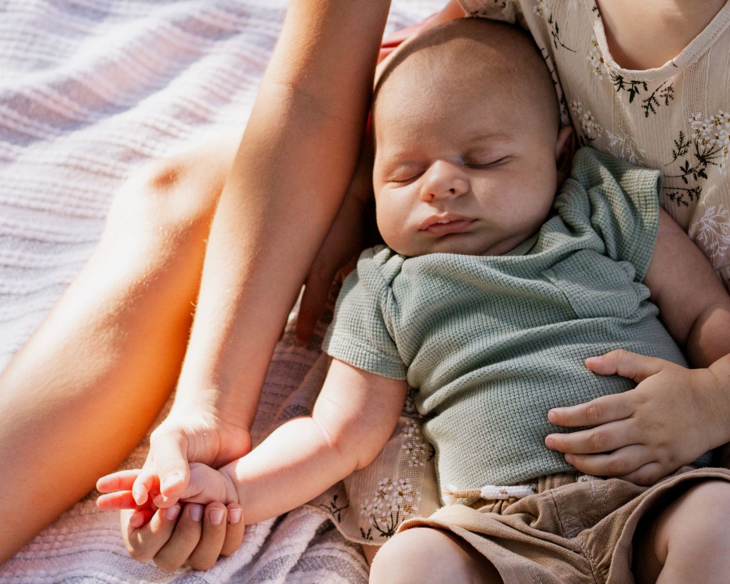 Sleeping newborn baby in green onesie cradled in mother's arms on a soft blanket in warm sunlight.