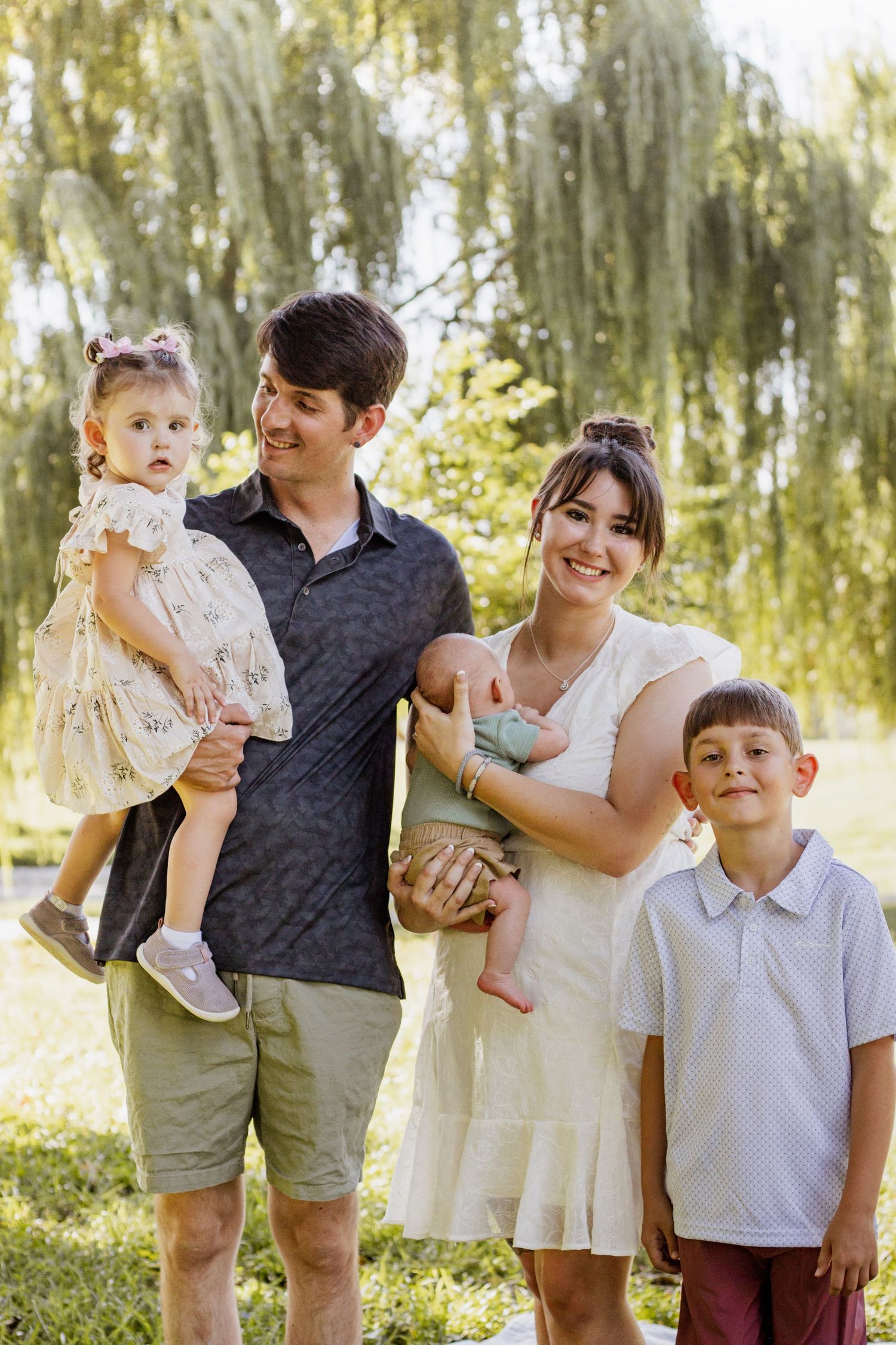 Happy family of five posing outdoors near a willow tree, parents holding three young children on a sunny day.