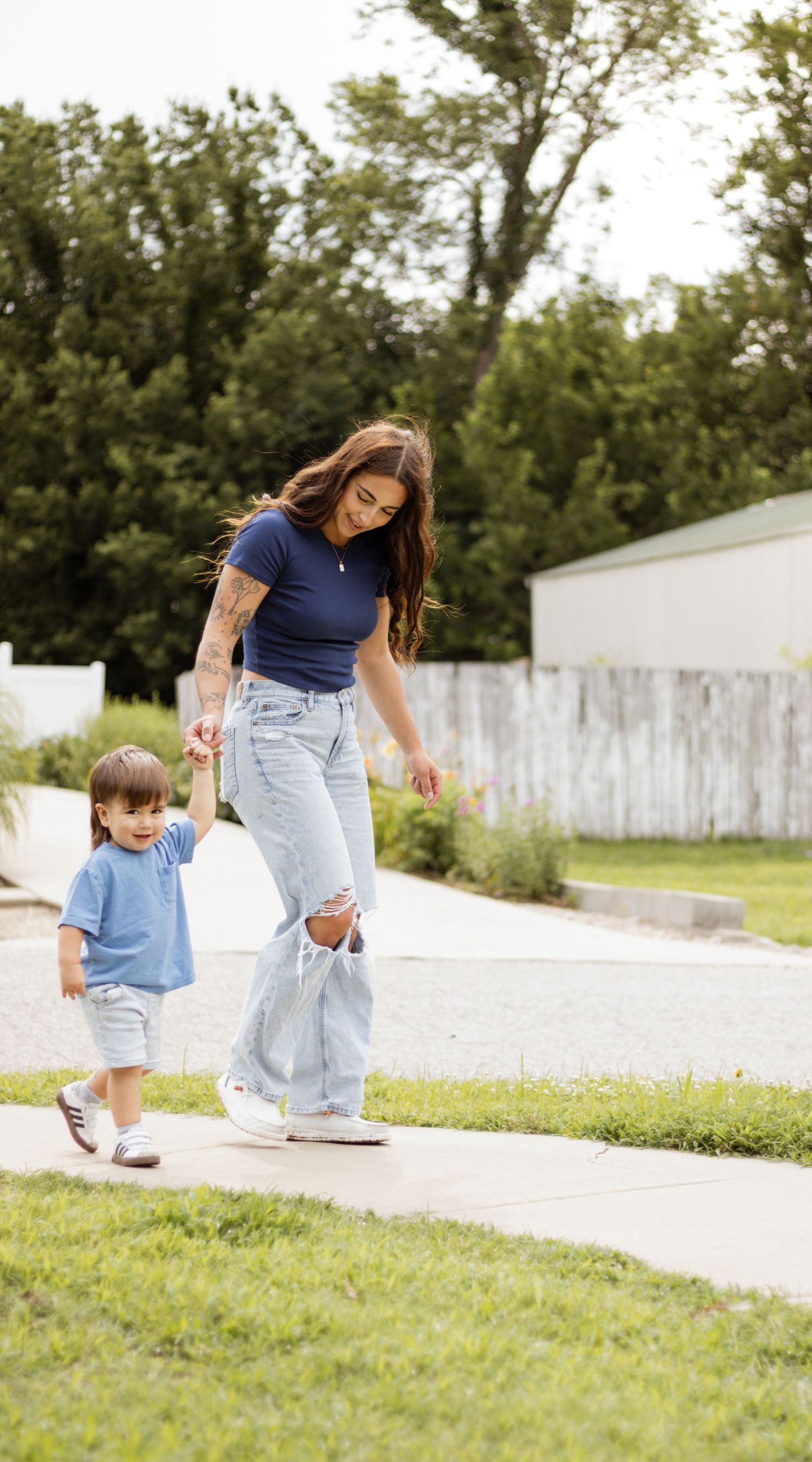 Young mother in blue outfit holding toddlers hand while walking on sunny suburban sidewalk.