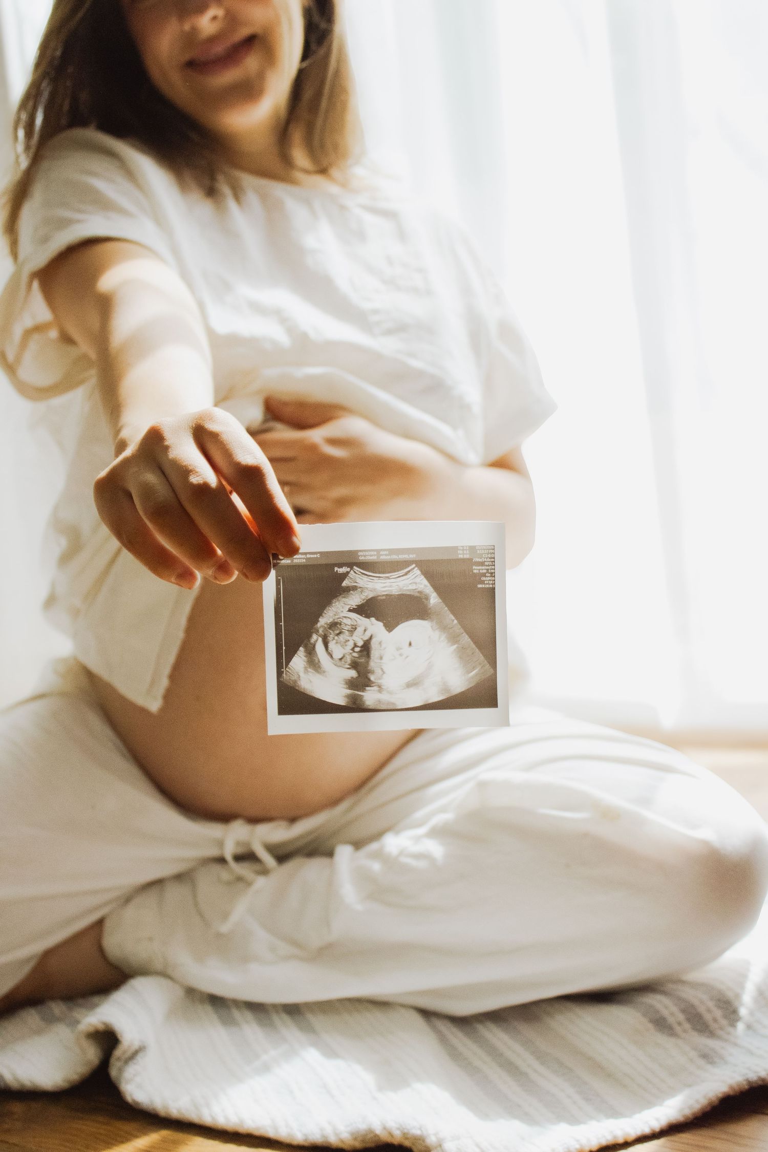 Pregnant woman in white linen outfit extending ultrasound photo toward camera showing baby scan image.