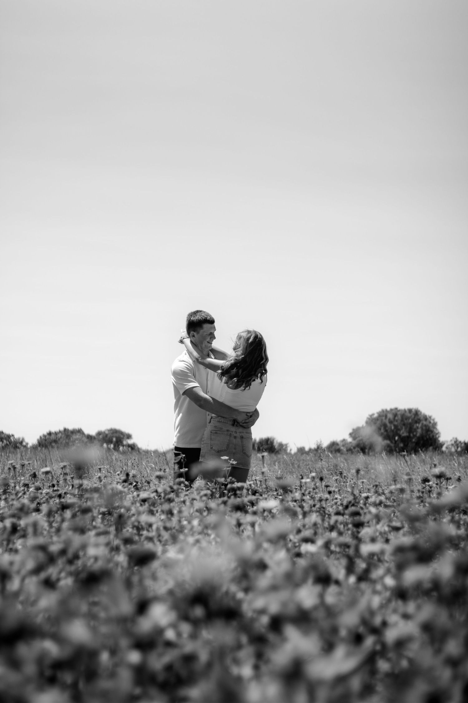Black and white photo of a romantic couple embracing in a blooming field, gazing at each other under a clear sky.