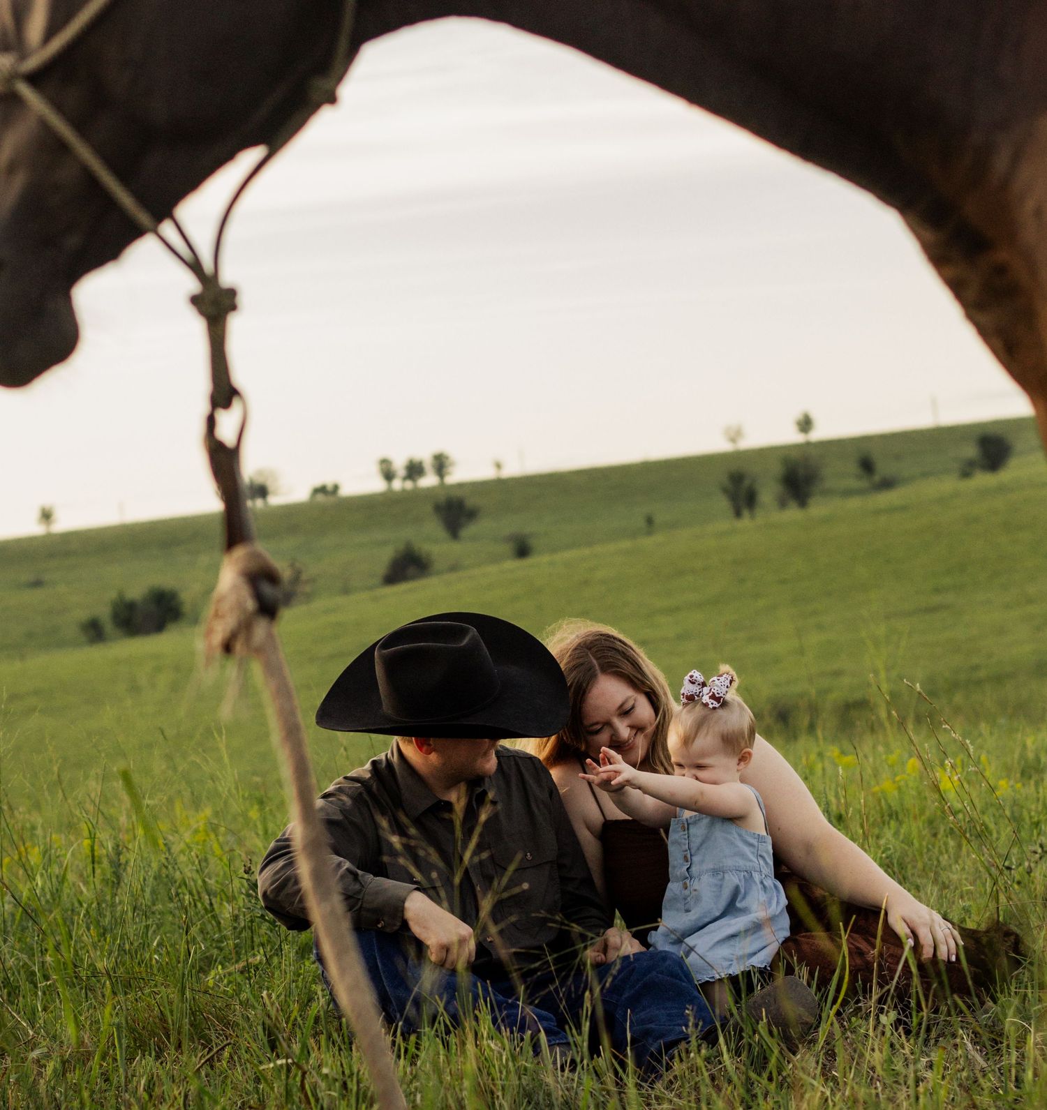 A couple sits in a green field with their baby, framed by a horse in the foreground on a farm.