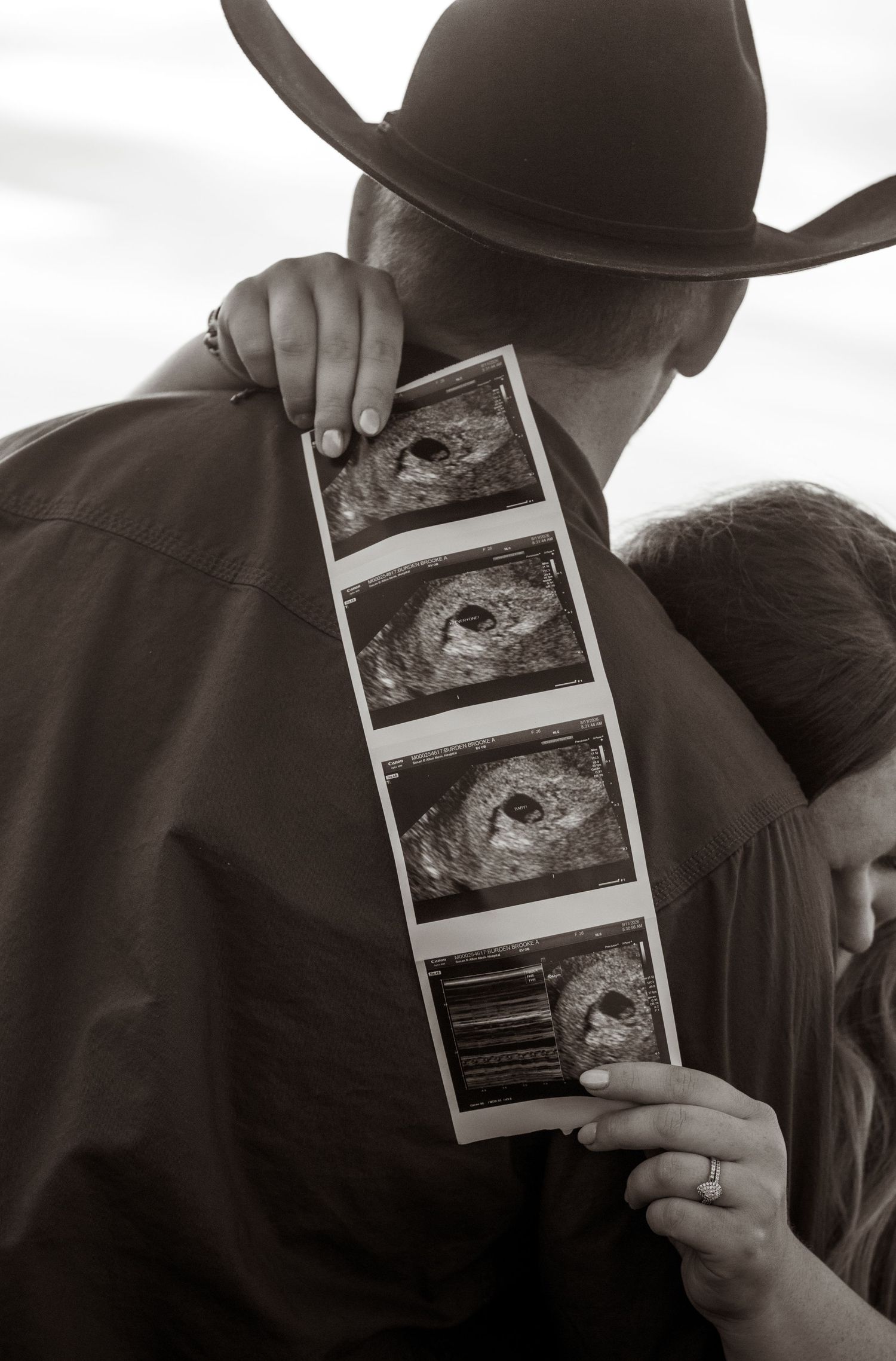Couple embracing while holding ultrasound photos, man wearing cowboy hat, black and white pregnancy announcement photo.